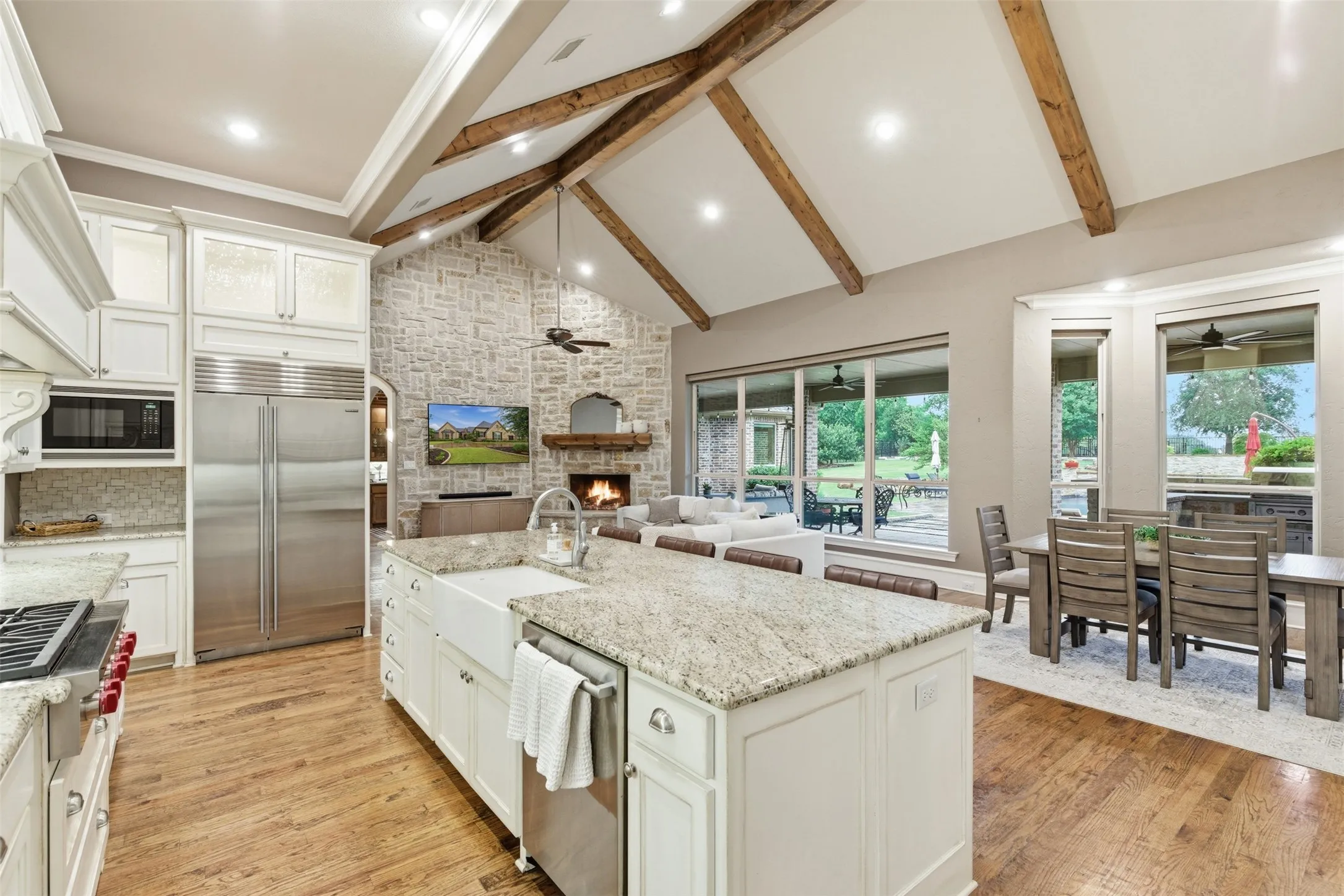 Expansive open-concept living area featuring a vaulted ceiling with exposed wood beams, a stone fireplace, wood-finish flooring, a kitchen island with a farmhouse sink, and a dining area with outdoor views