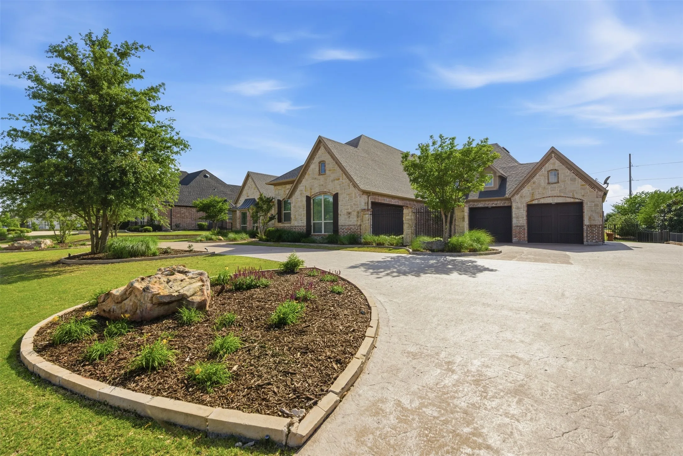 Expansive concrete driveway leading to a stone and brick exterior home featuring multiple garage doors, gabled rooflines, and mature landscaping