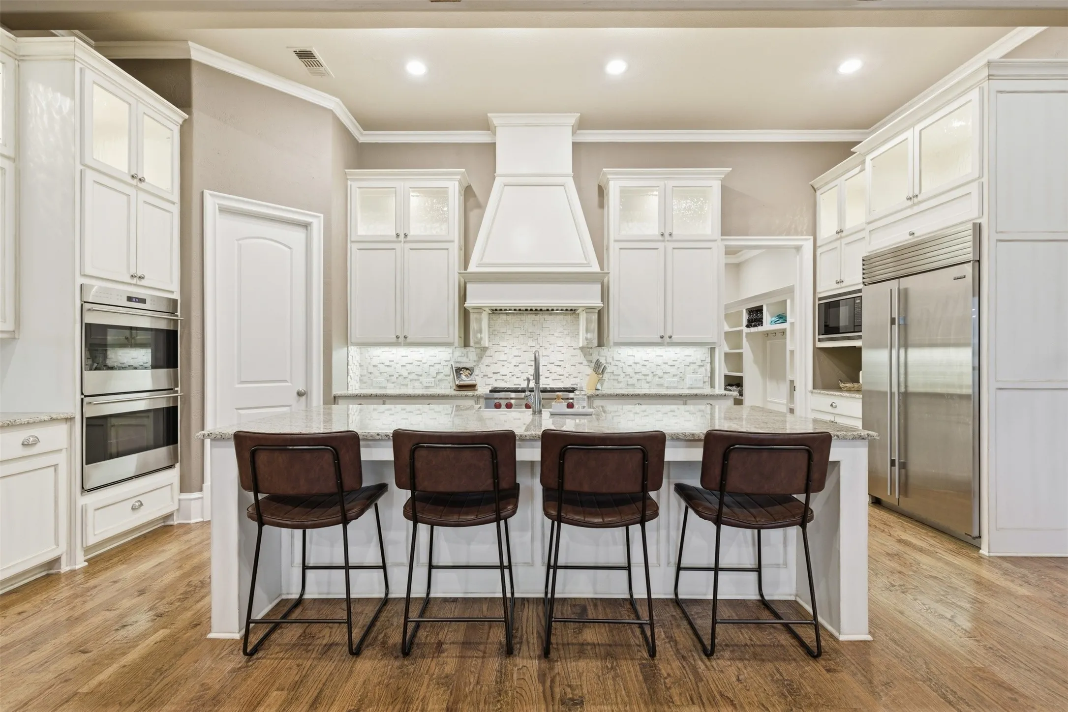 Central island with seating for four, surrounded by white cabinetry with upper glass-front cabinets