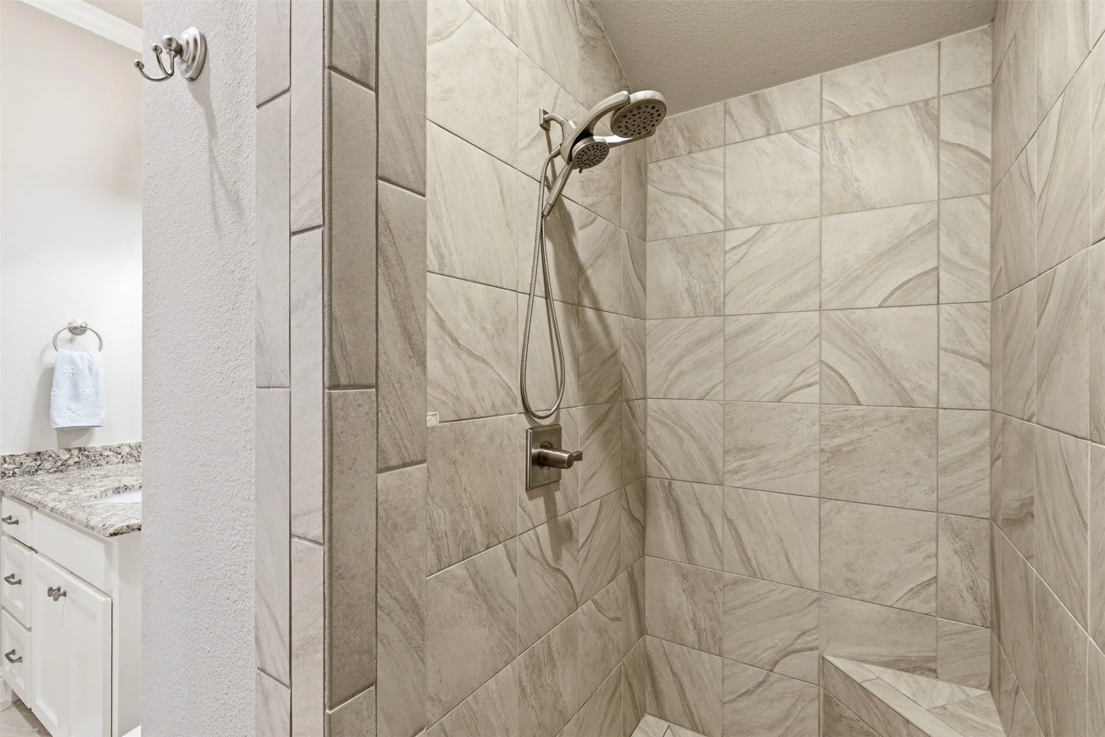 Walk-in shower featuring light-toned tile with a subtle pattern, dual shower heads, and an integrated corner bench