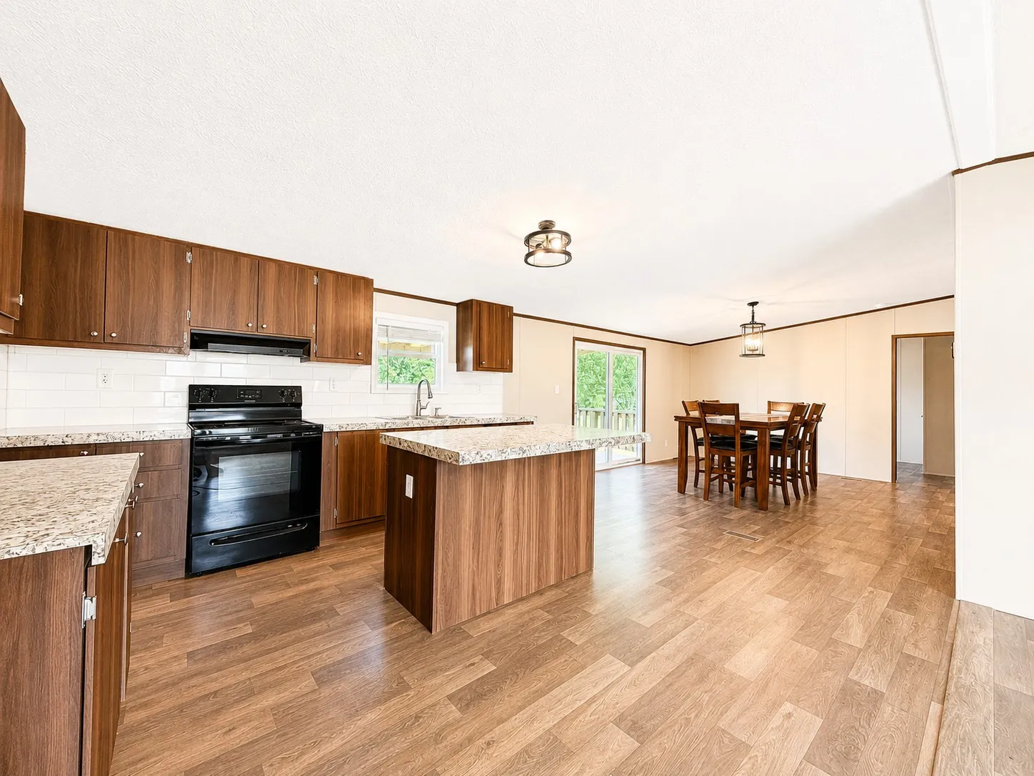 Open-concept kitchen and dining area featuring wood-finish flooring, wood cabinetry, light-toned granite-style countertops, and a subway tile backsplash