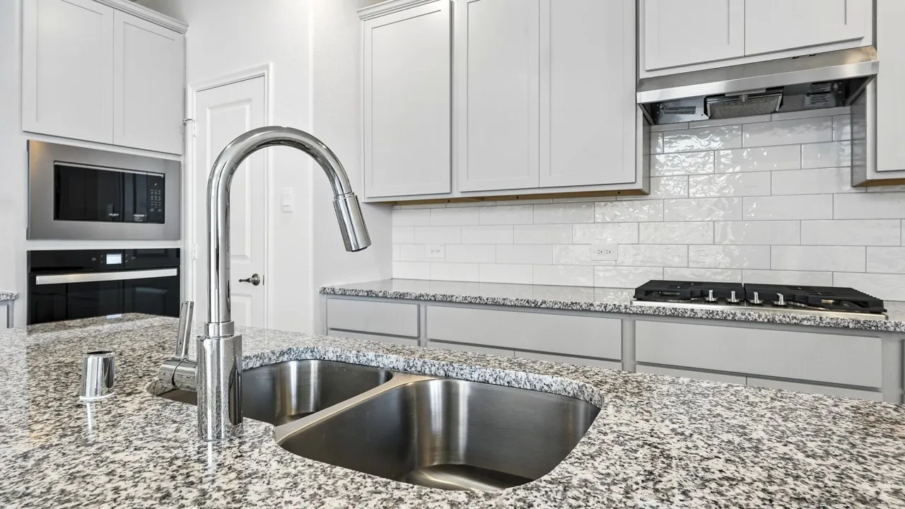 Kitchen featuring a double basin stainless steel sink with a chrome gooseneck faucet, granite countertops, white subway tile backsplash, white cabinetry, and stainless steel appliances including a built-in oven and microwave