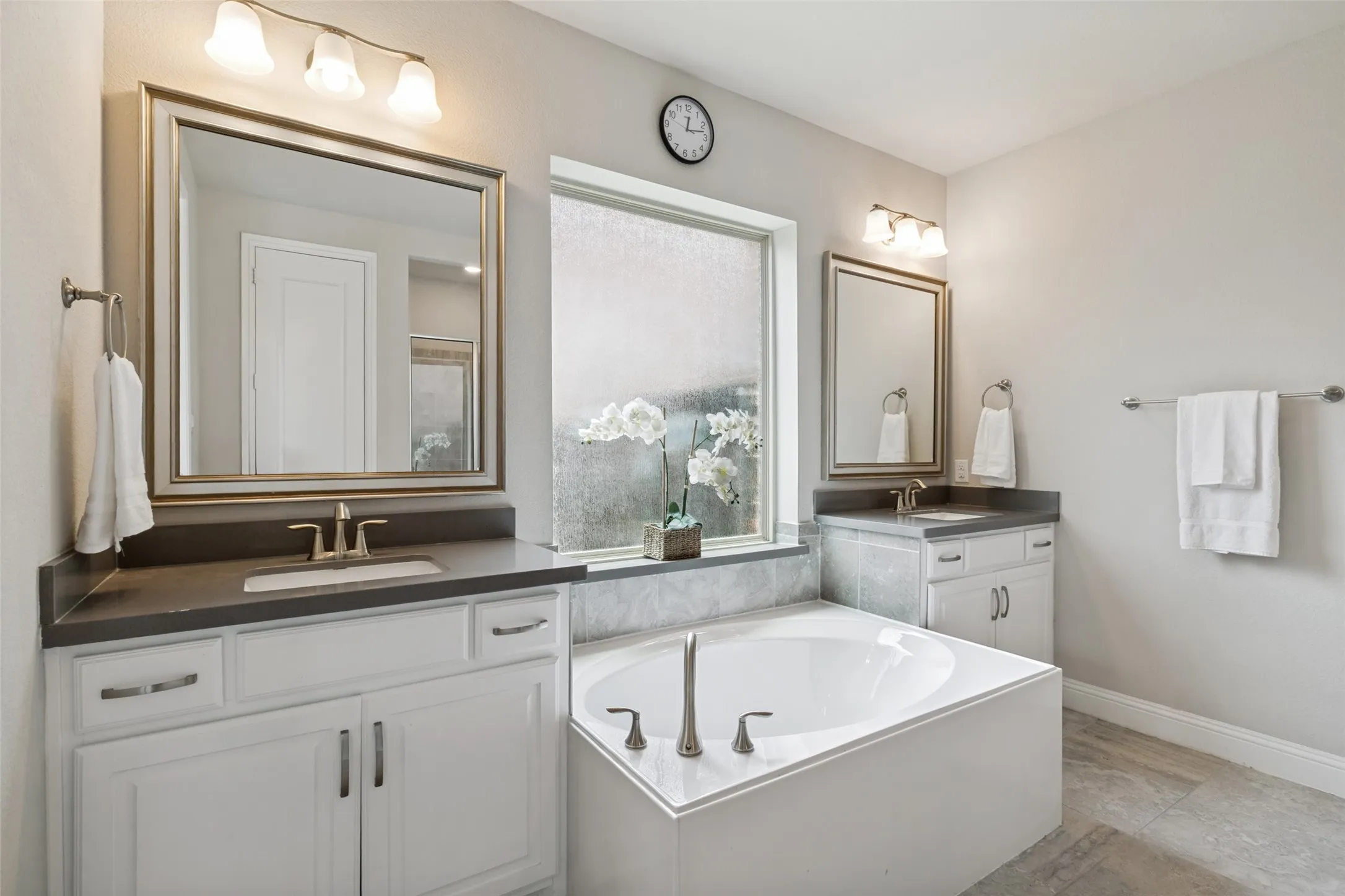 Bathroom featuring a built-in soaking tub, dual vanity sinks with dark countertops, and light-toned tile flooring