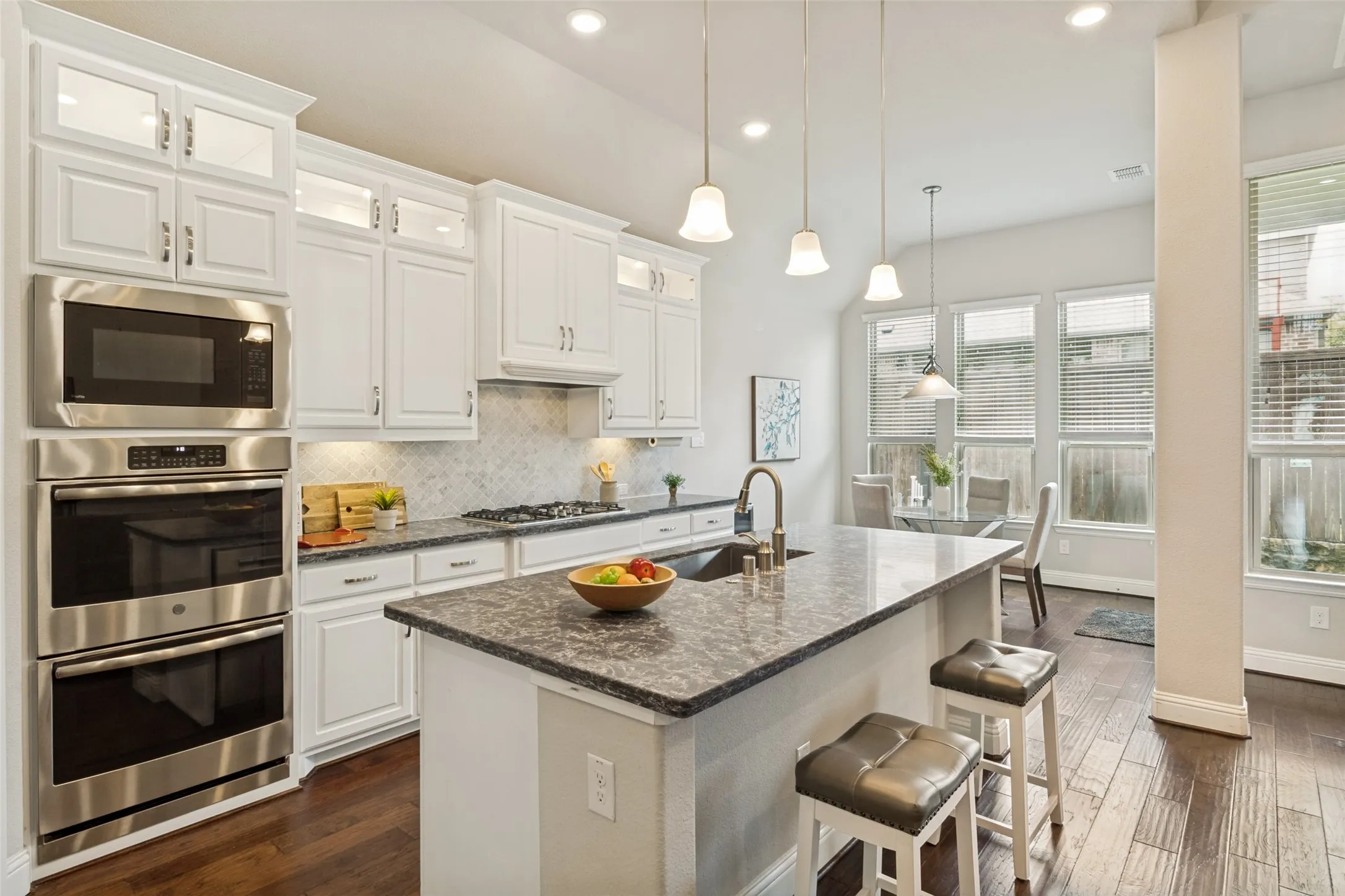 Kitchen featuring a central island with stone countertop, stainless steel appliances including a double oven and microwave, white cabinetry with glass-front uppers, a tile backsplash, and wood-finish flooring