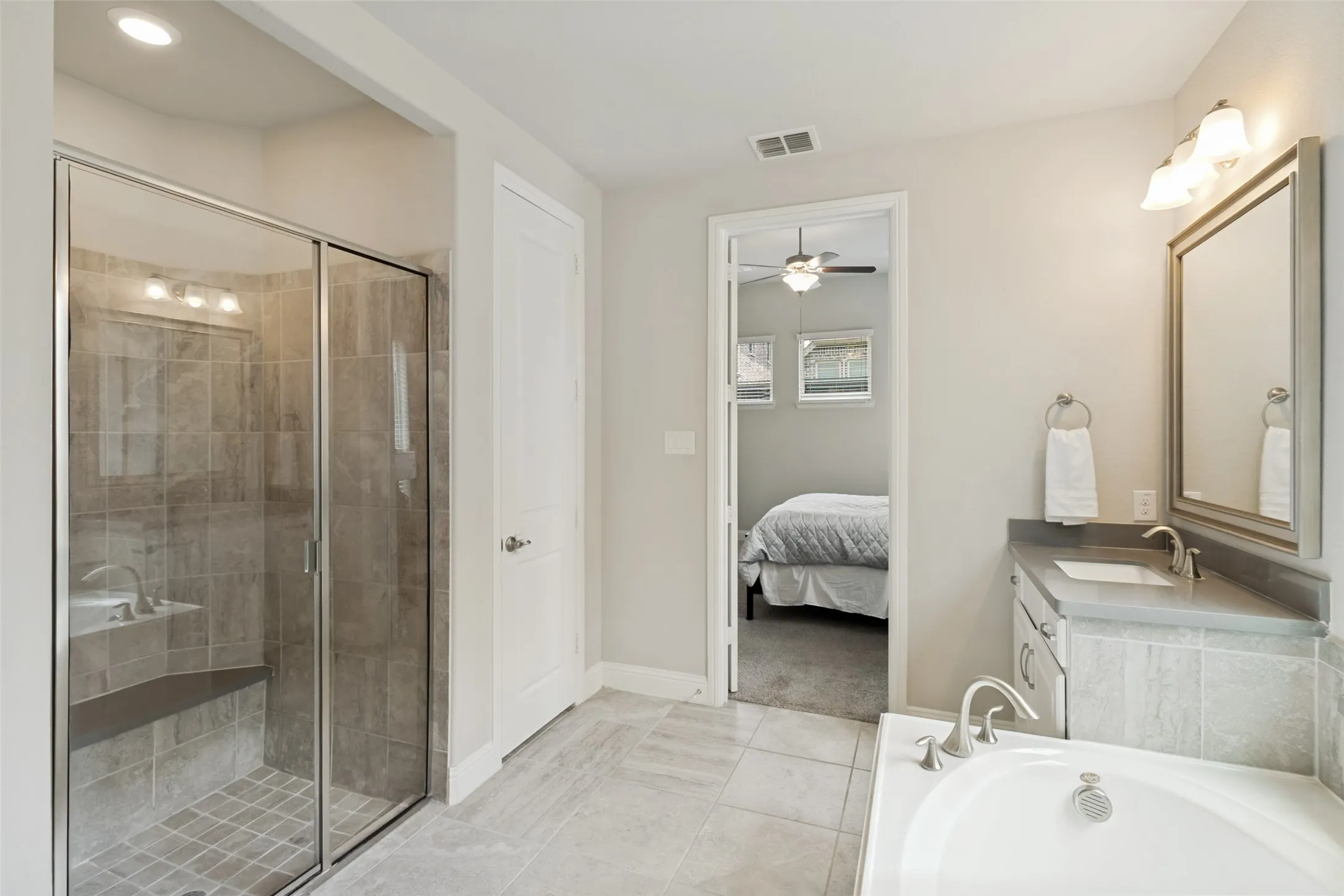 Bathroom featuring a glass-enclosed shower with tiled walls and a built-in bench, a separate soaking tub, a vanity with an integrated sink and mirror, and light-toned tile flooring
