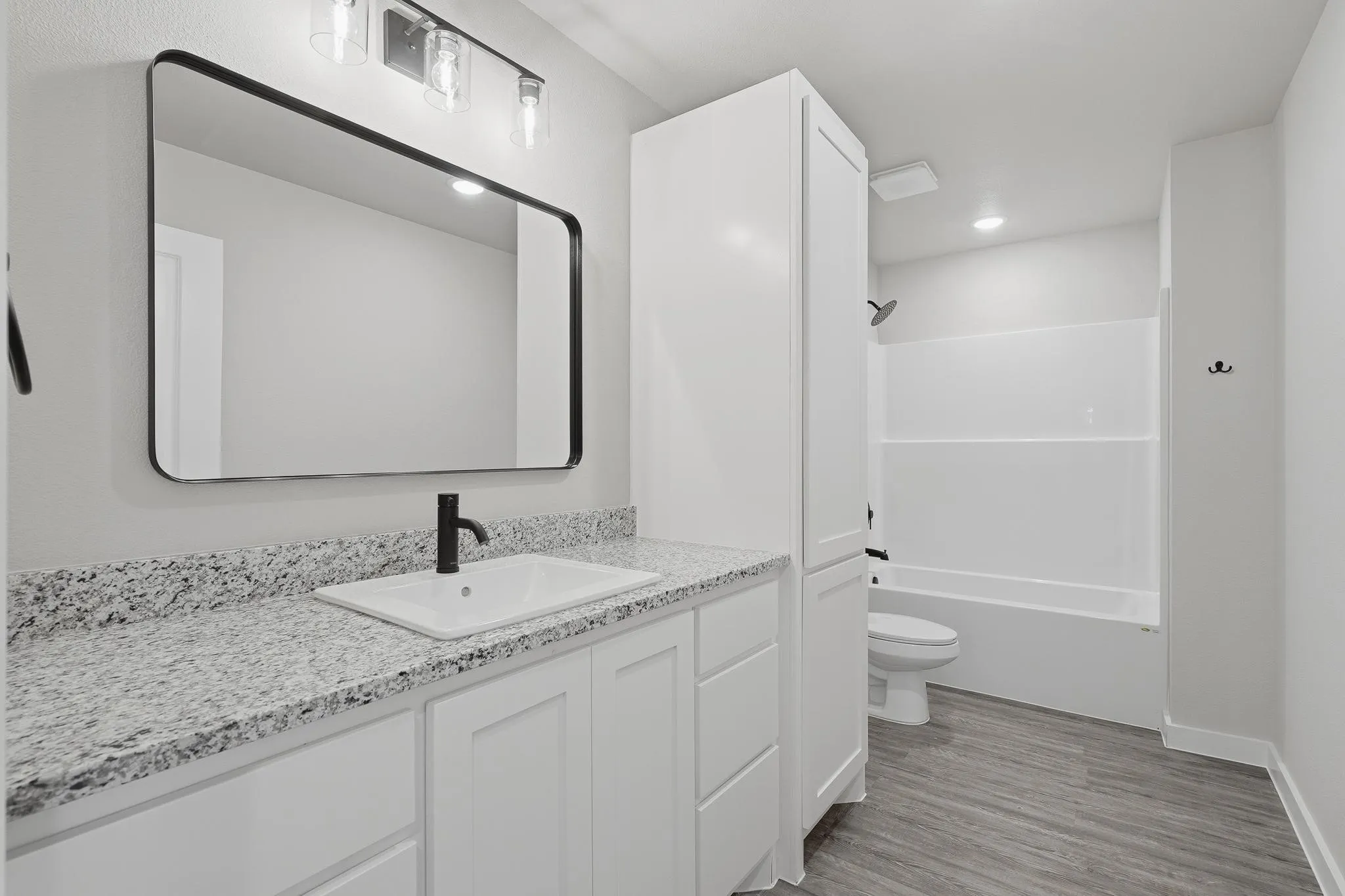 Modern bathroom featuring a rectangular vanity mirror, granite-look countertop, white cabinetry with ample storage, a shower-tub combination, and wood-finish flooring