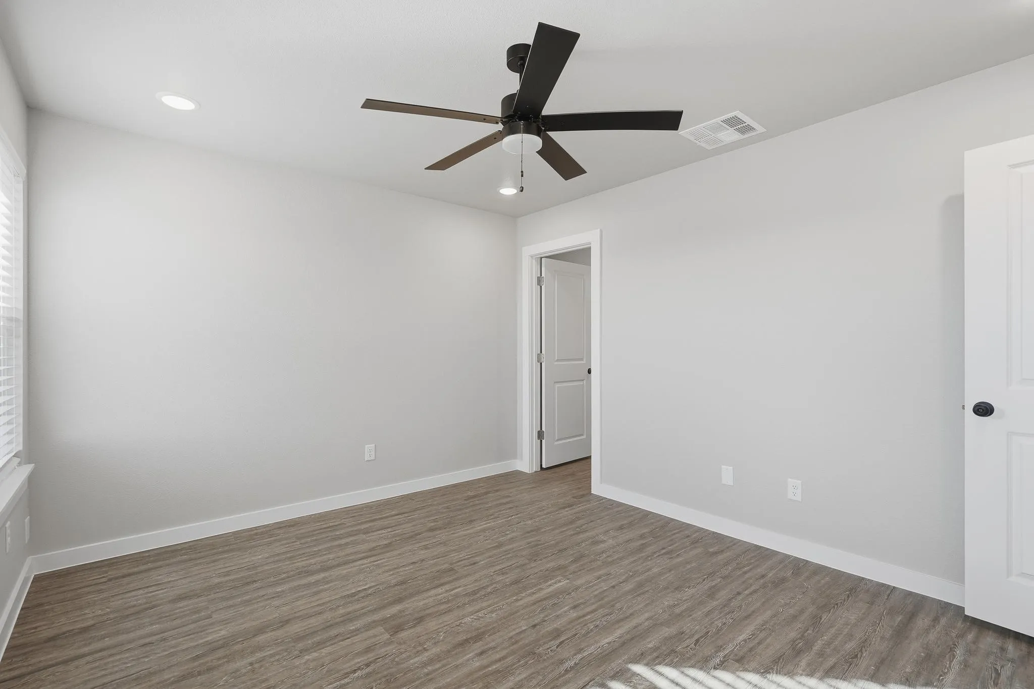 Spacious room featuring wood-finish flooring, light gray walls, white baseboards, and a contemporary black ceiling fan with integrated lighting