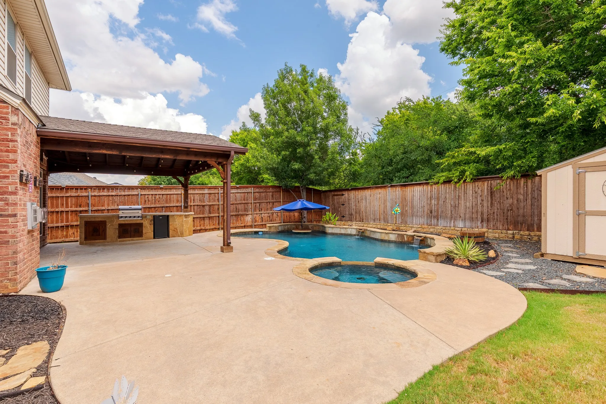 Expansive concrete patio featuring a built-in outdoor kitchen with grill, a covered pergola, a multi-level swimming pool, and an integrated spa