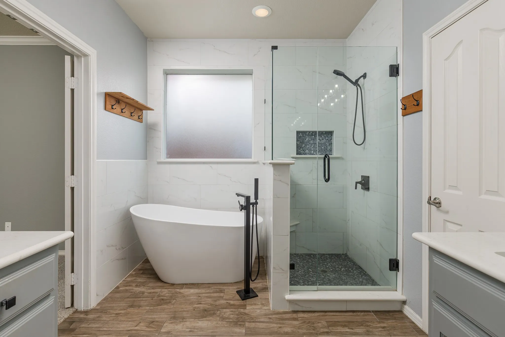 Contemporary bathroom featuring a freestanding tub with a matte black floor-mounted faucet, a frameless glass shower enclosure with a built-in bench and recessed shelf, wood-finish flooring, and light gray wall paint