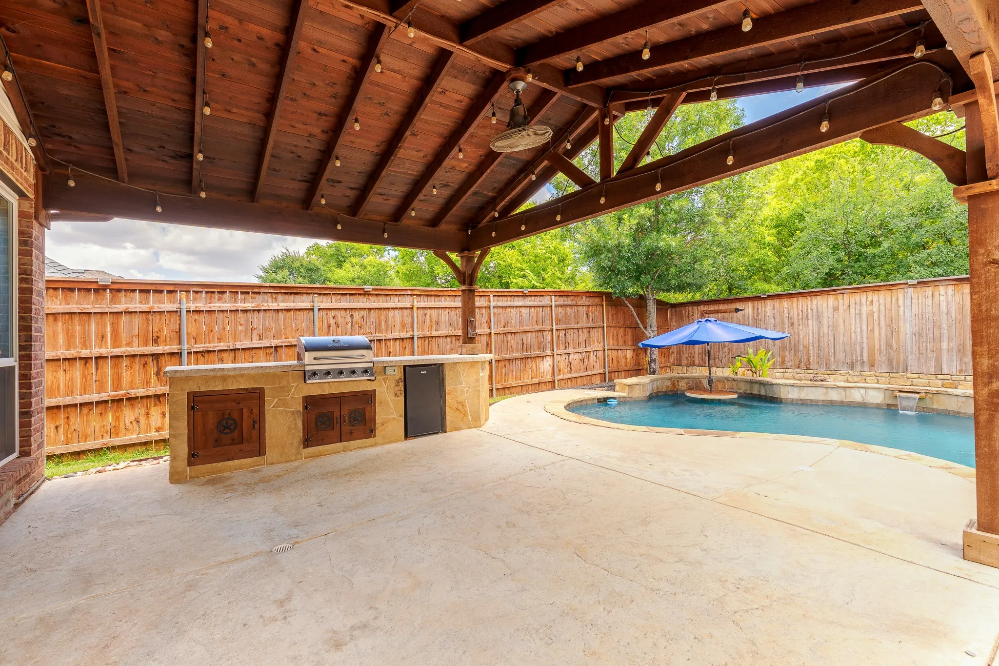 Outdoor living area featuring a covered patio with exposed wood ceiling, built-in outdoor kitchen with grill and refrigerator, and a freeform swimming pool with stone coping and water features