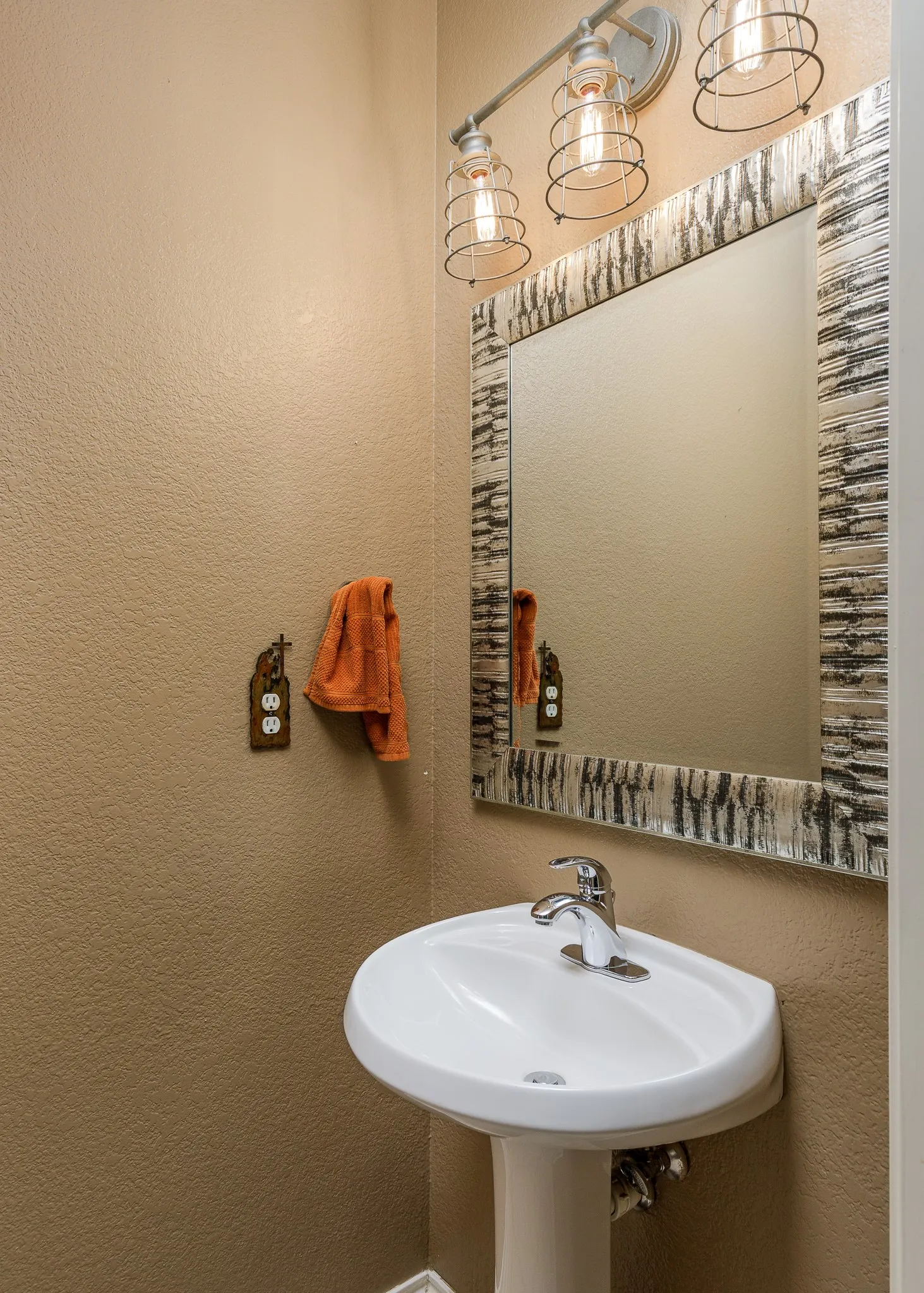 Pedestal sink with chrome faucet, complemented by a decorative framed mirror and a three-light vanity fixture featuring caged bulbs