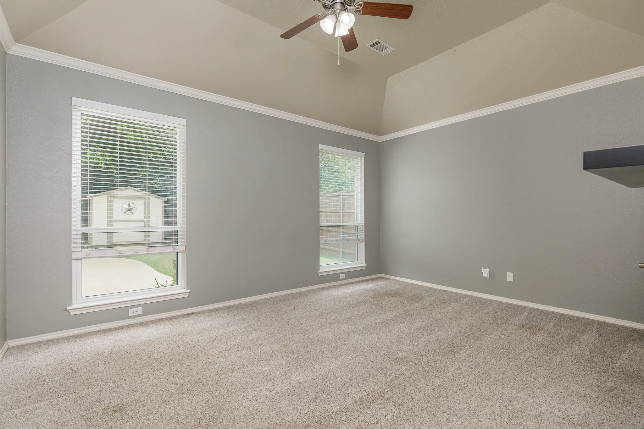 Carpeted interior space featuring vaulted ceilings with a ceiling fan, two windows with blinds, crown molding, and a partial dark-toned built-in shelf