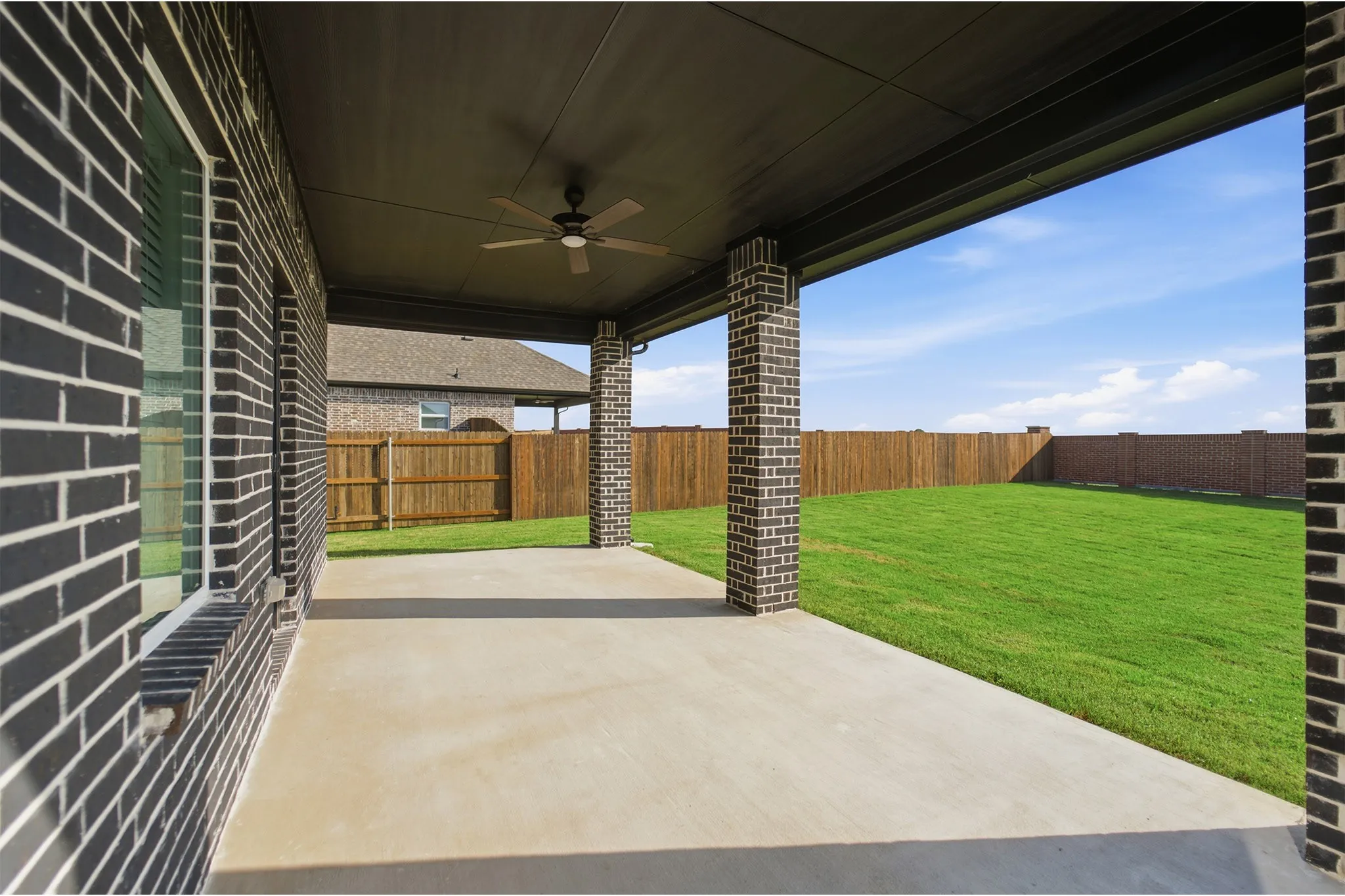 Covered patio featuring a ceiling fan, concrete flooring, brick columns, and a dark soffit, opening to a grass lawn and wood fence