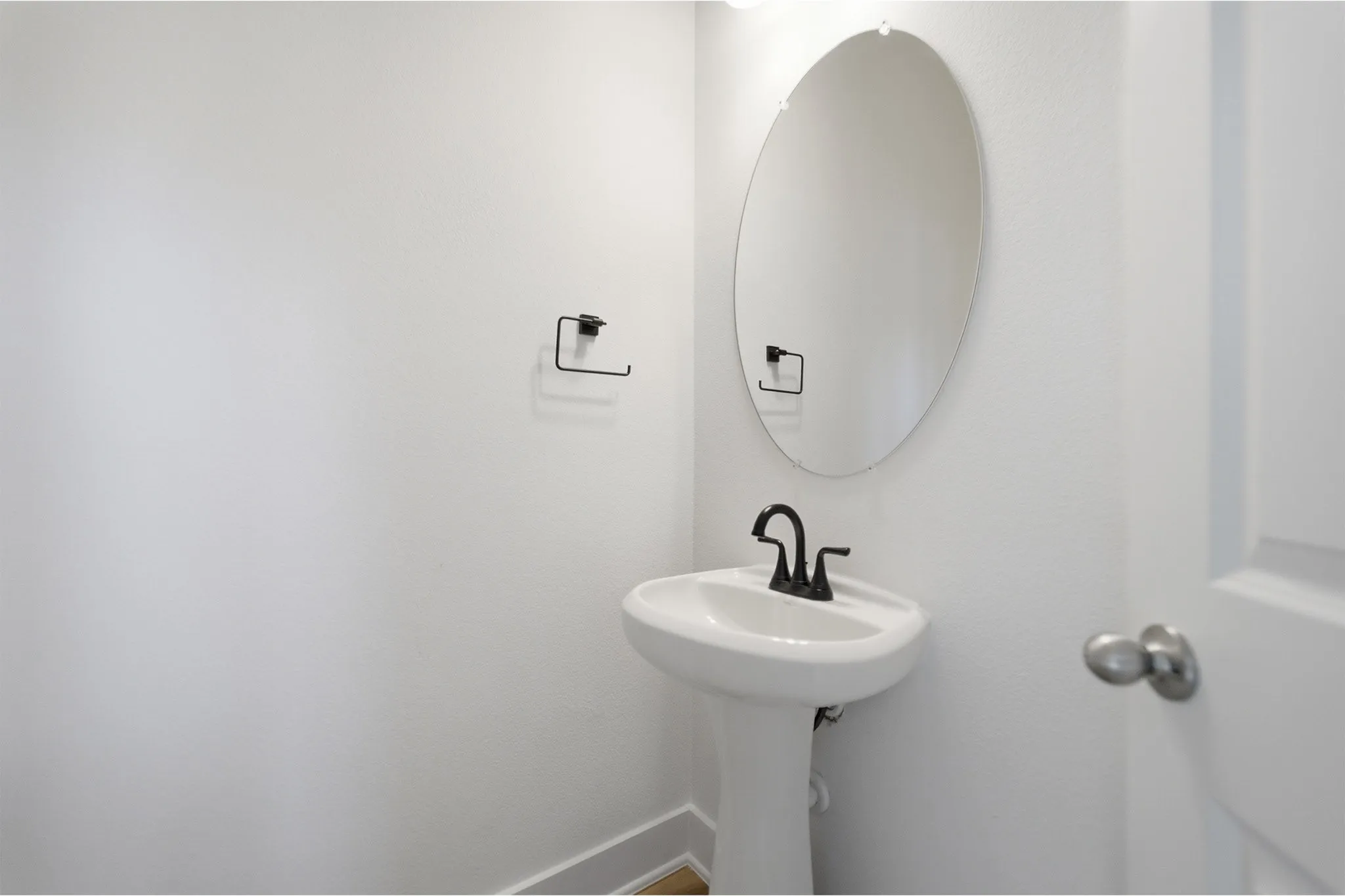 Pedestal sink with matte black faucet, wall-mounted oval mirror, and light wood-finish flooring