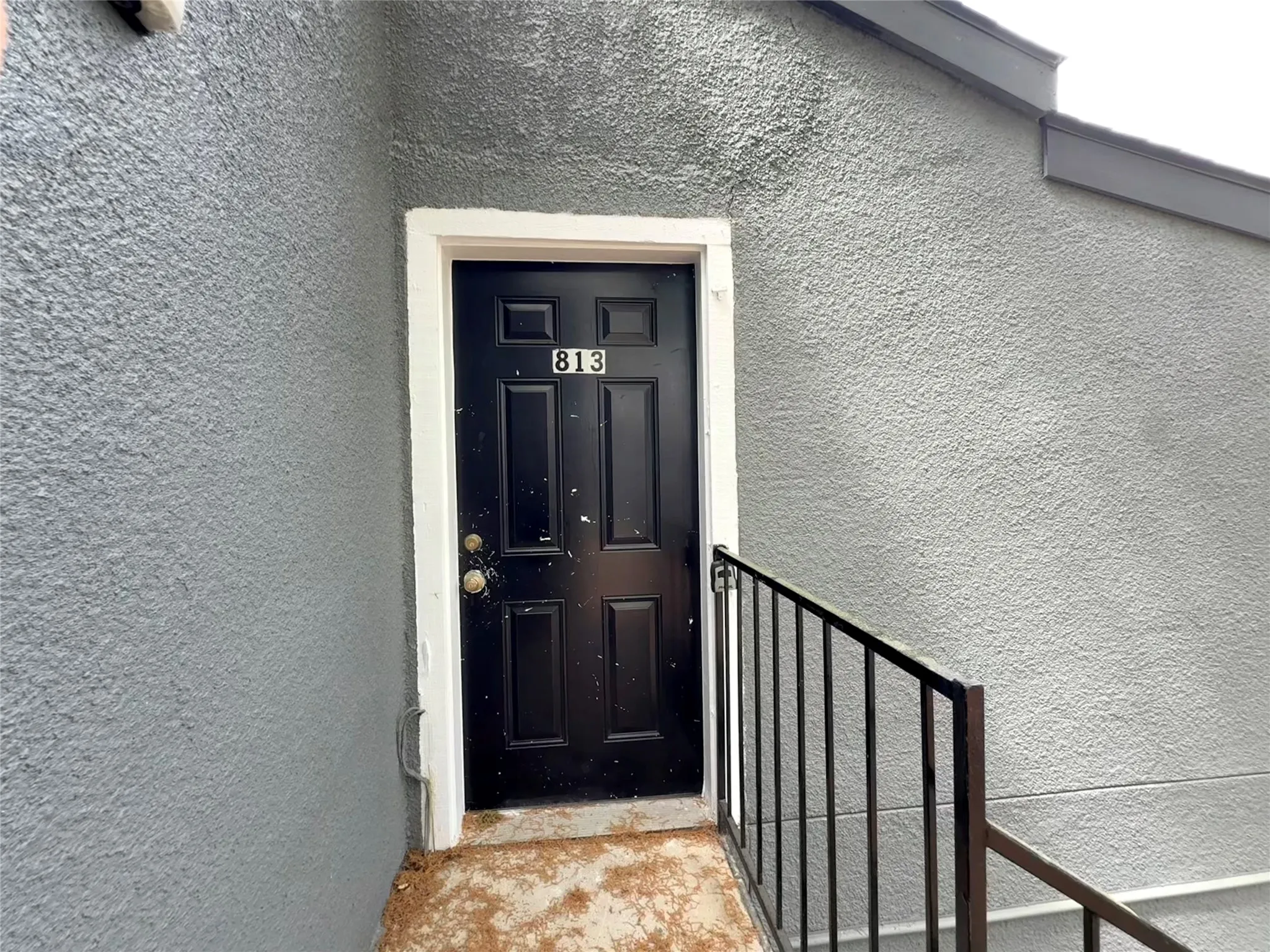 Black paneled entry door with white trim, featuring a metal railing and stucco exterior finish
