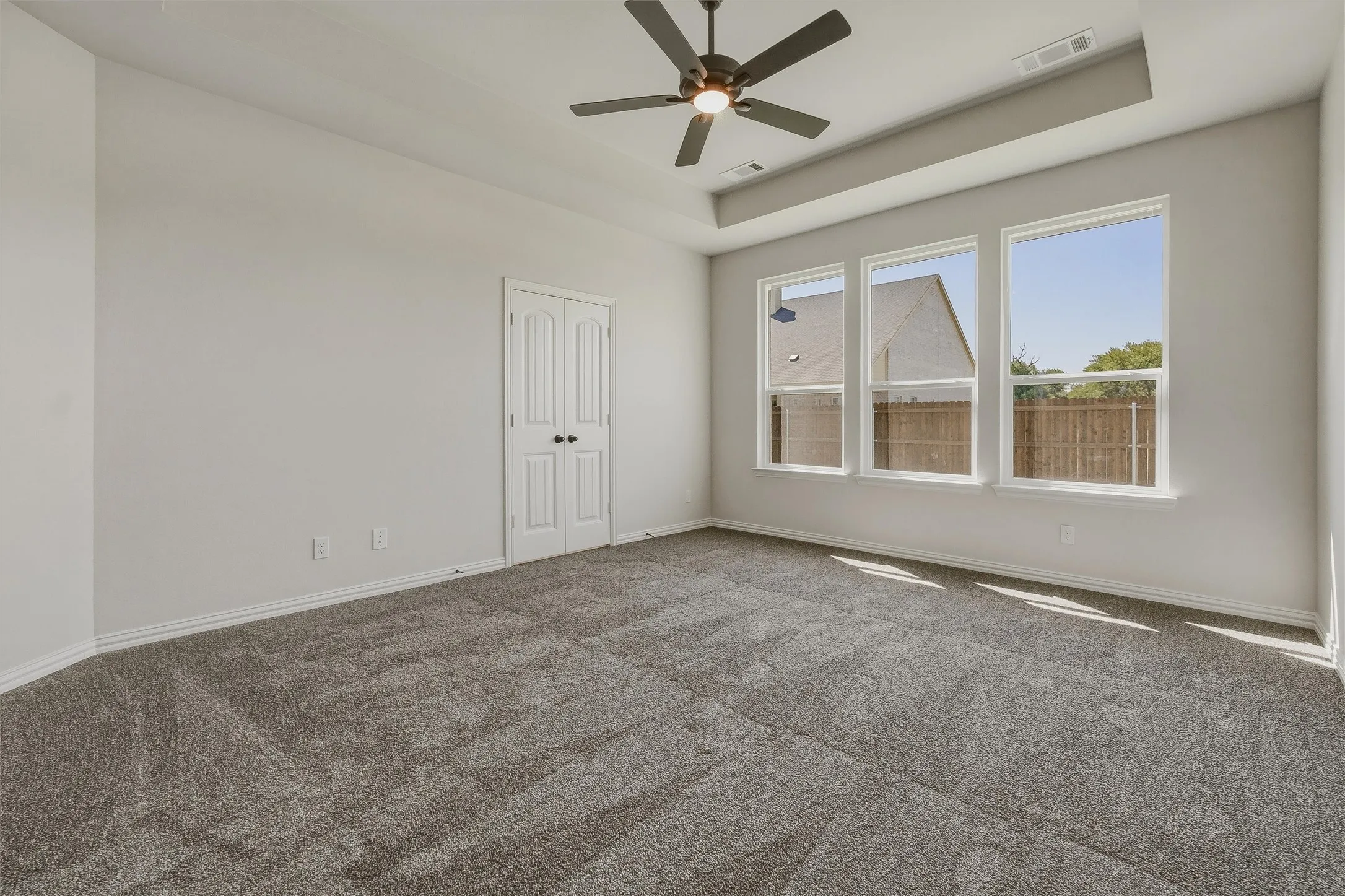 Recessed ceiling detail with an attached ceiling fan, three expansive windows, neutral wall tones, and gray carpeting
