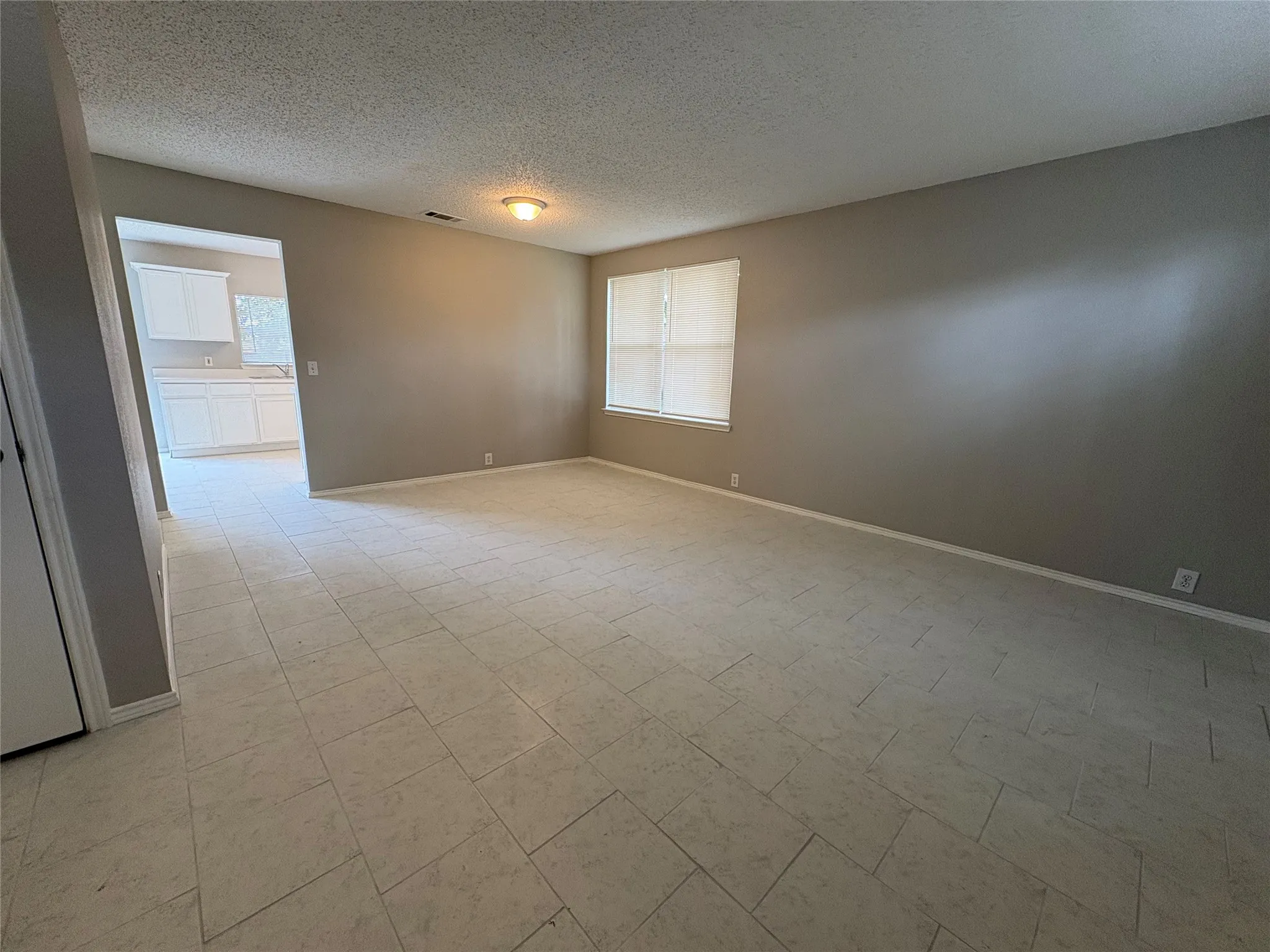 Spacious living area featuring light-toned tile flooring, neutral wall paint, and a large window with blinds