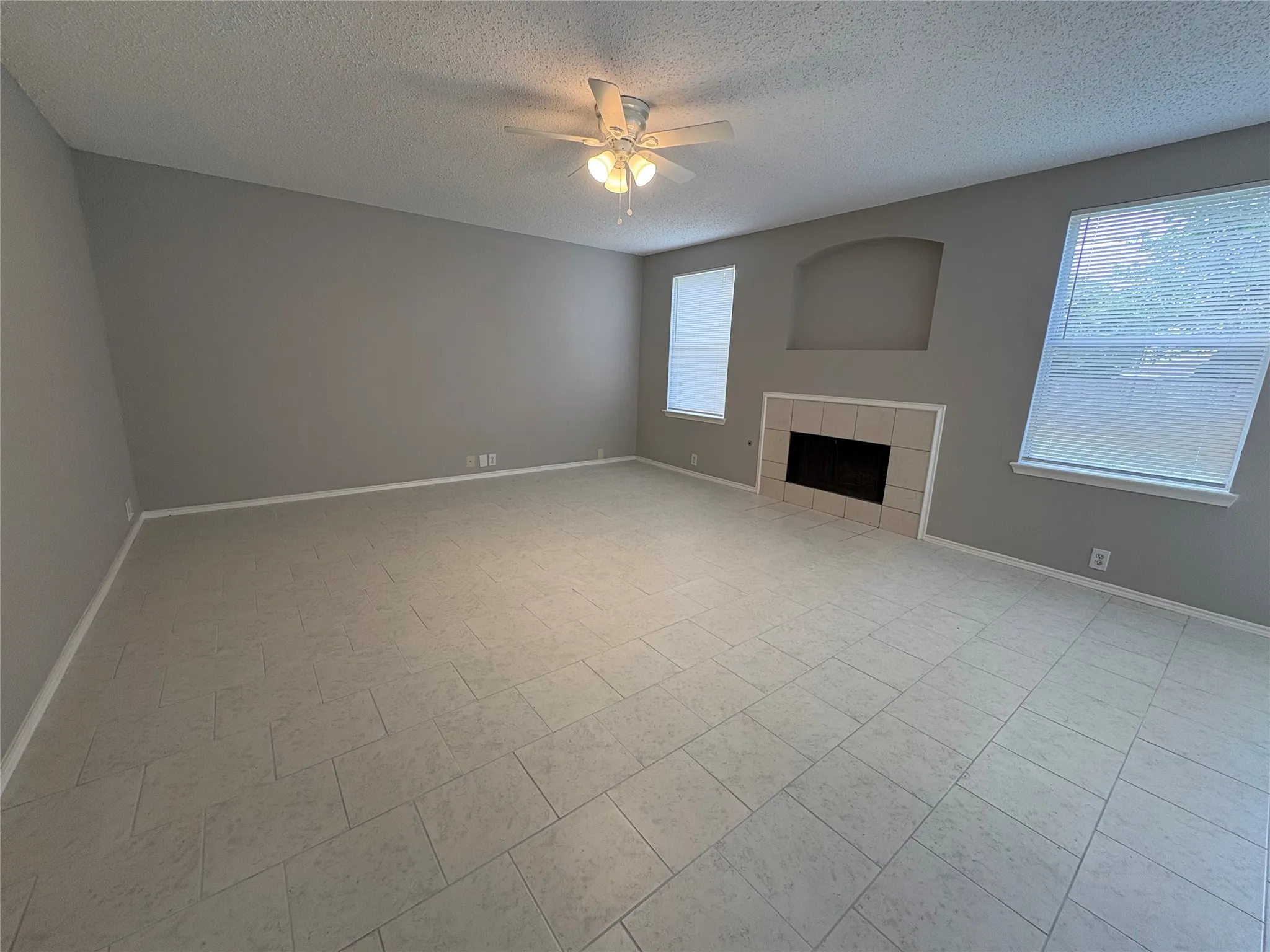 Spacious living area featuring neutral-toned tile flooring, a built-in fireplace with a tiled surround, two windows with blinds, a ceiling fan with integrated lighting, and an arched wall niche