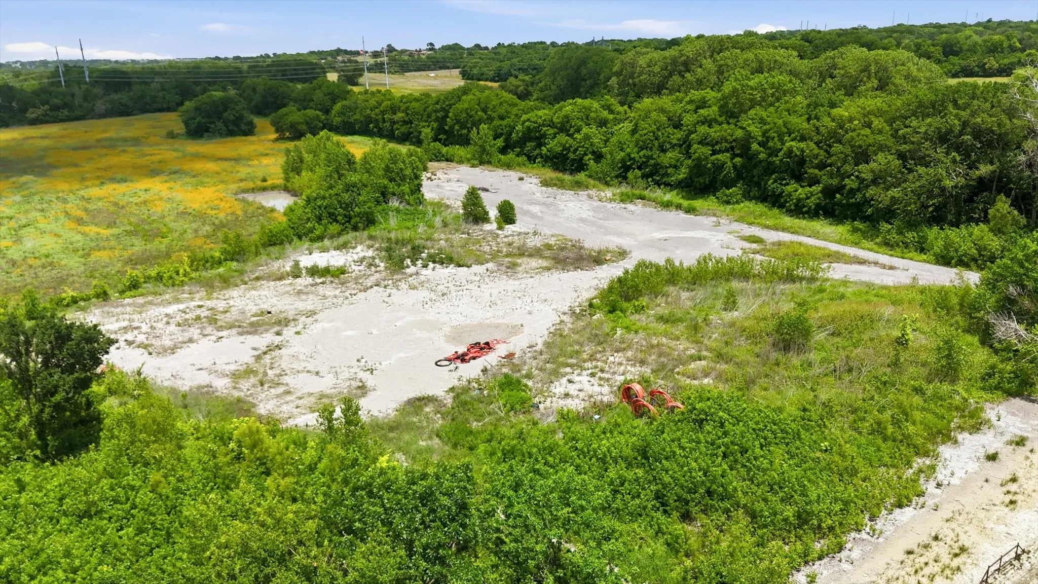 Expansive land parcel featuring a gravel-surfaced area, diverse natural vegetation, and a backdrop of mature trees