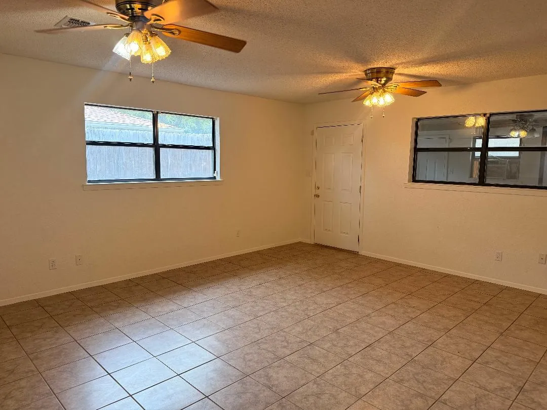 Spacious interior room featuring neutral-toned tile flooring, two ceiling fans with integrated lighting, a white paneled door, and two windows with dark frames