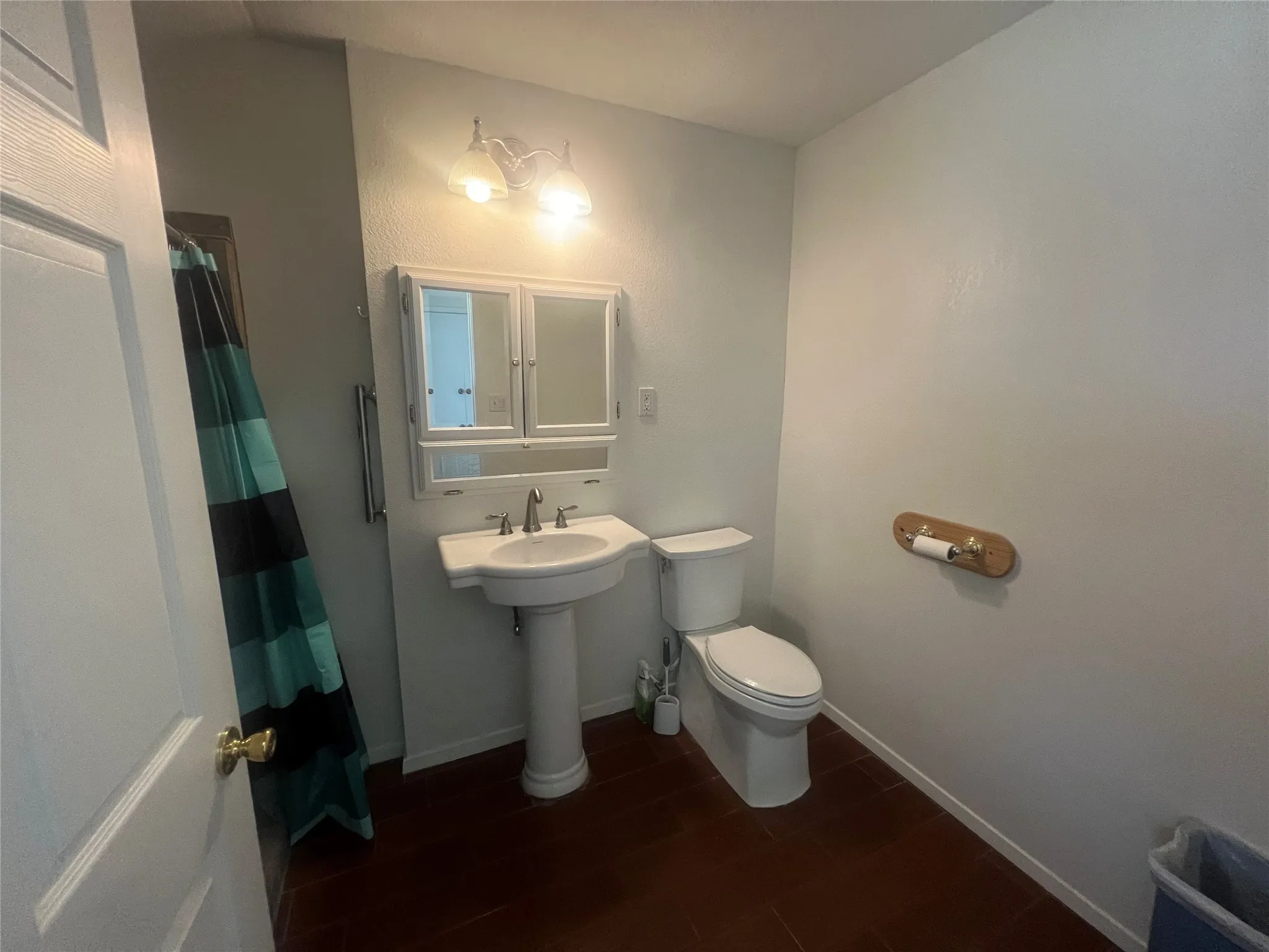 Pedestal sink with chrome fixtures and overhead vanity lighting, white two-door mirrored medicine cabinet, white toilet, and dark wood-finish flooring