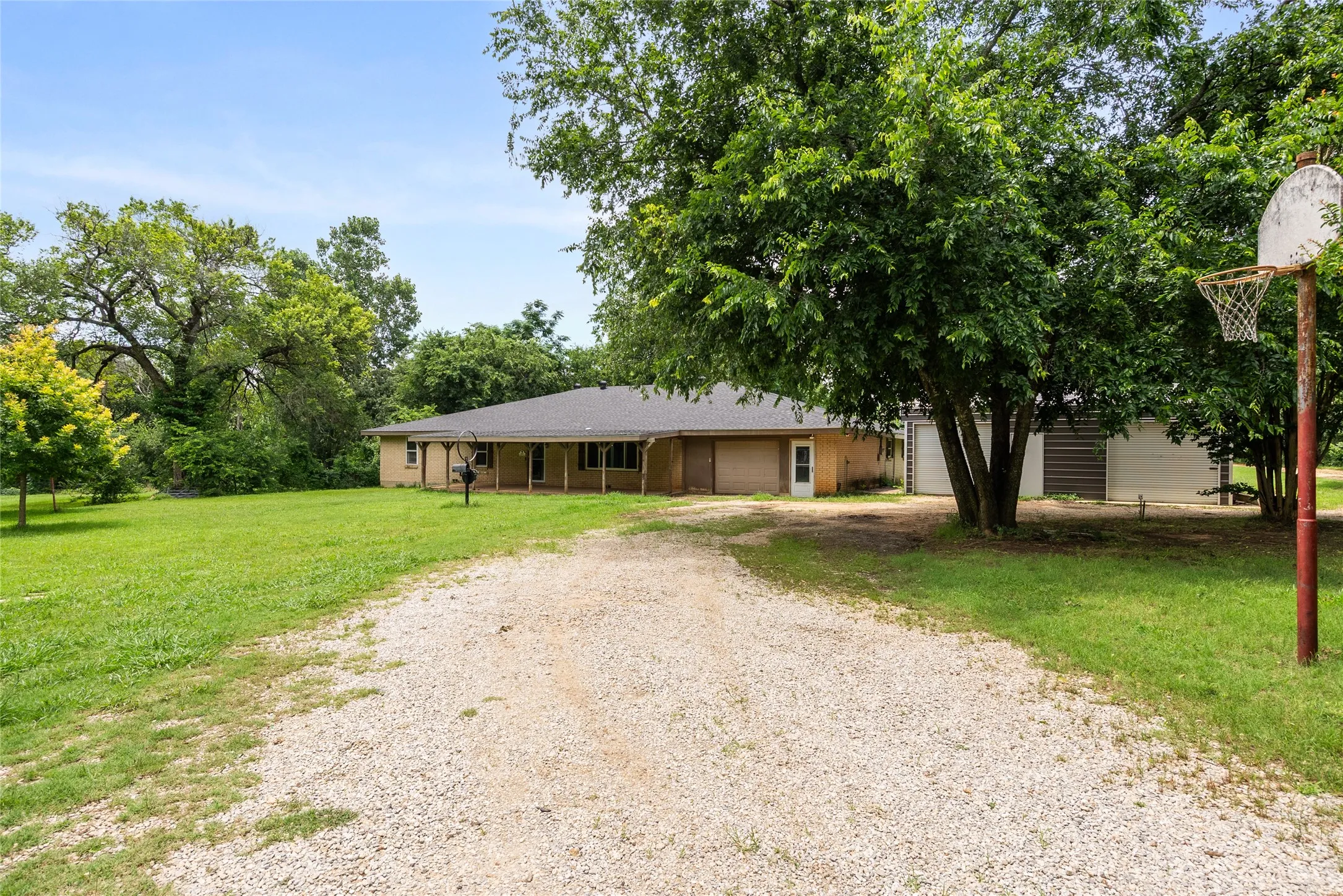 Ranch-style exterior with a covered front porch, gravel driveway, and surrounding mature trees