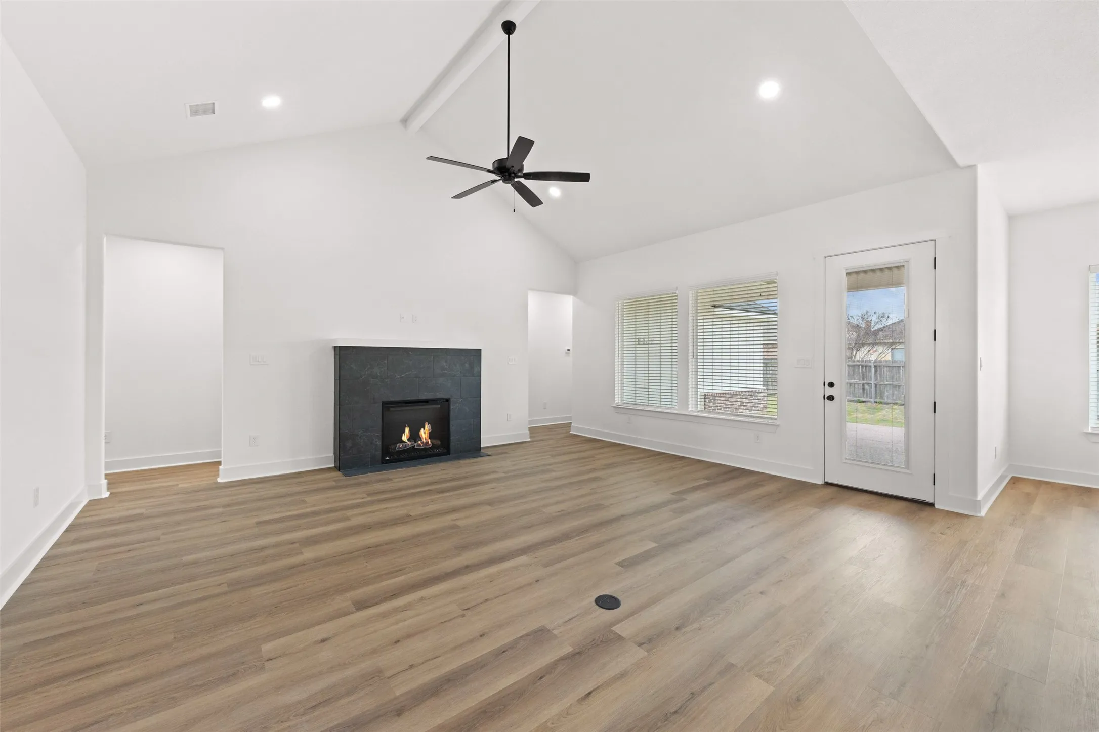 Spacious living area featuring vaulted ceilings, wood-finish flooring, a contemporary fireplace with a dark stone surround, a ceiling fan with integrated lighting, and a glass-paned exterior door