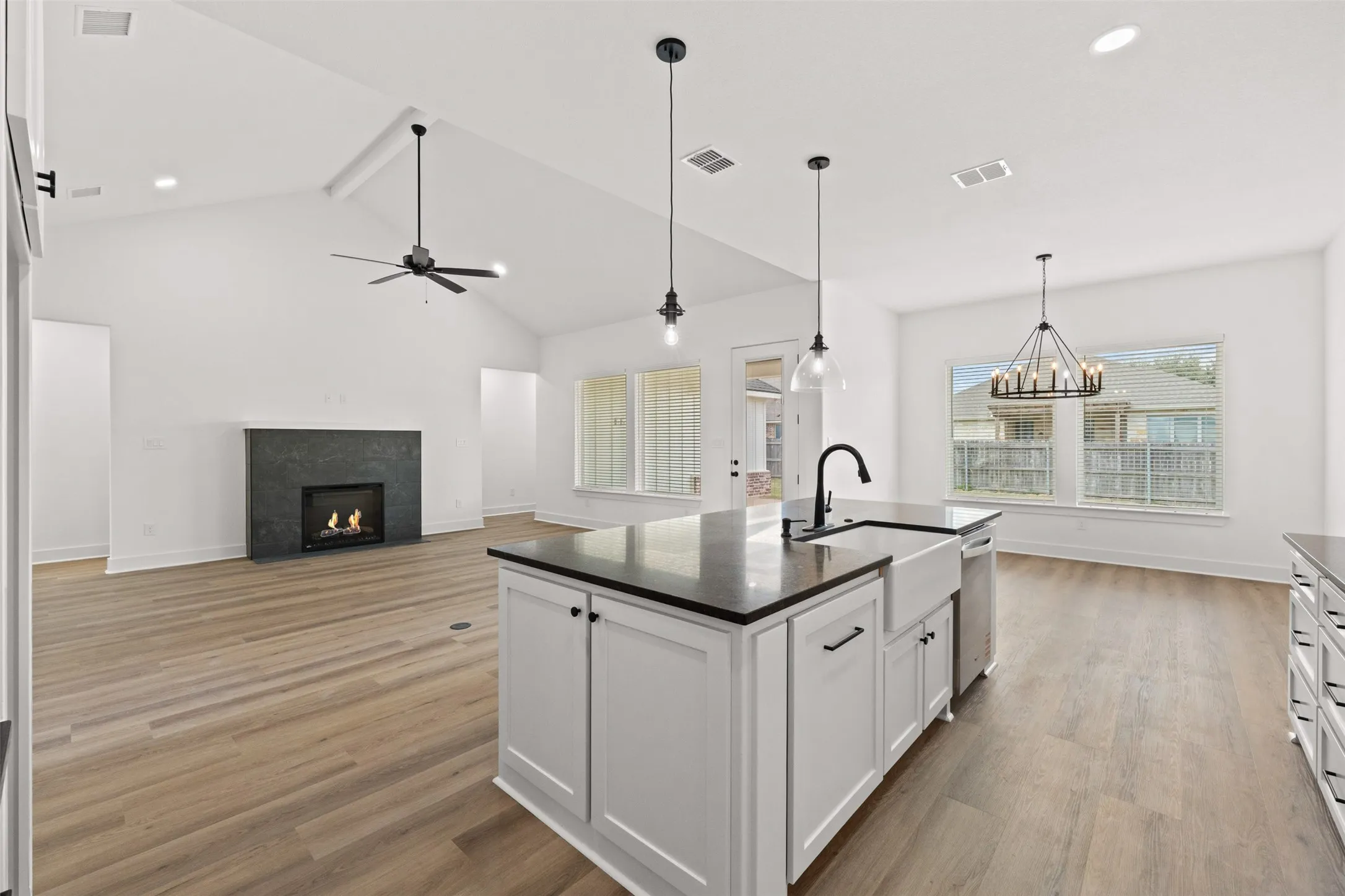 Open-concept interior featuring light wood-finish flooring, a kitchen island with a farmhouse sink and black countertop, a fireplace with a dark stone surround, vaulted ceilings, and multiple windows for natural light