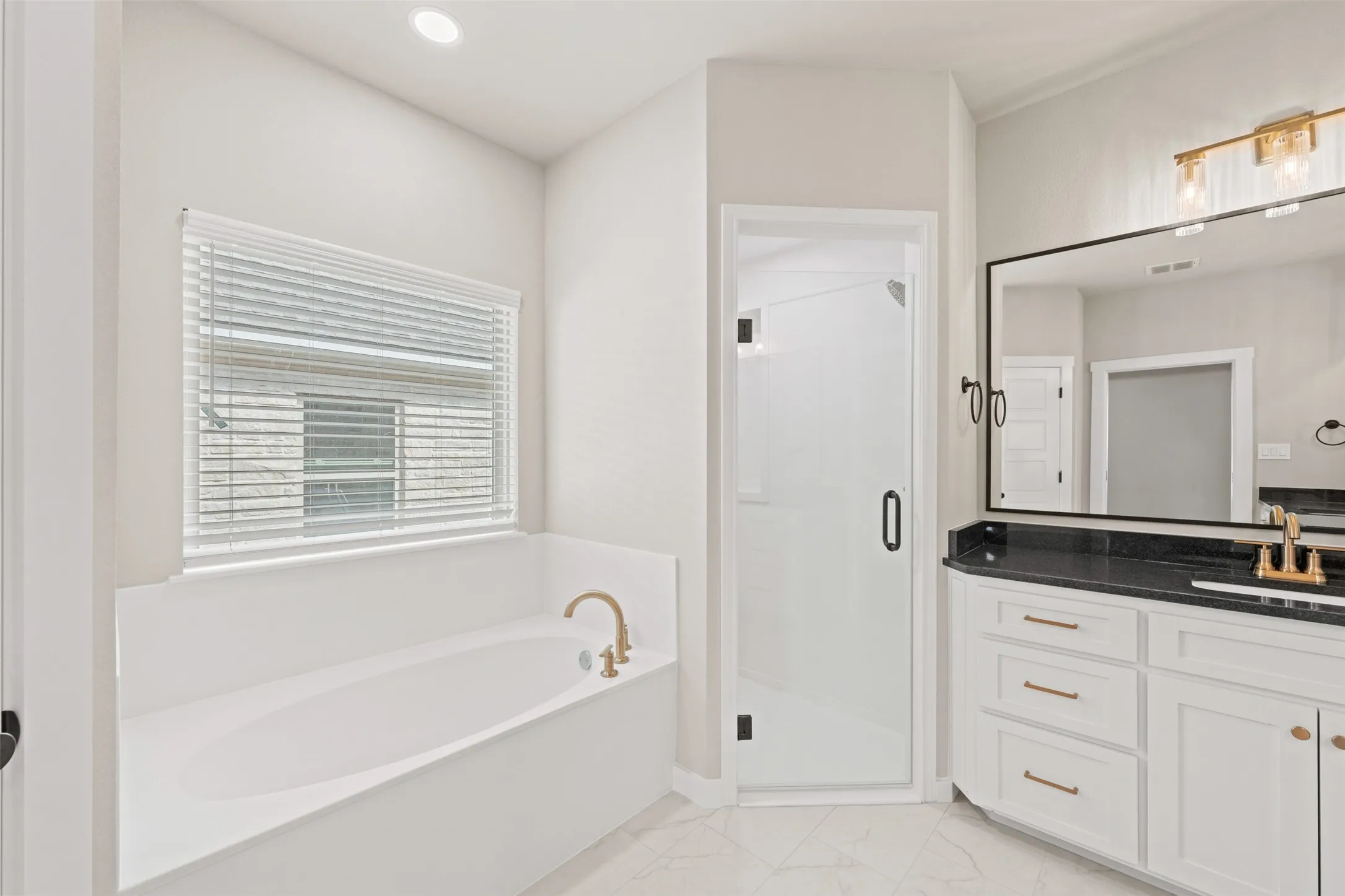 Bathroom featuring a built-in soaking tub with brass fixtures, a walk-in shower with a frameless glass door, a double vanity with a dark countertop and gold-toned hardware, and a large wall-mounted mirror
