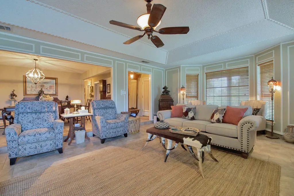 Spacious living area featuring tile flooring, a tray ceiling with a fan, and an expansive bay window with blinds