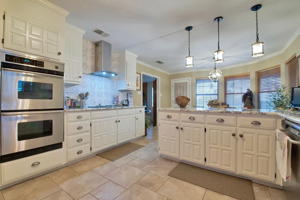 Kitchen featuring white paneled cabinetry, stainless steel double ovens, a stainless steel range hood, stone countertops, and tile flooring