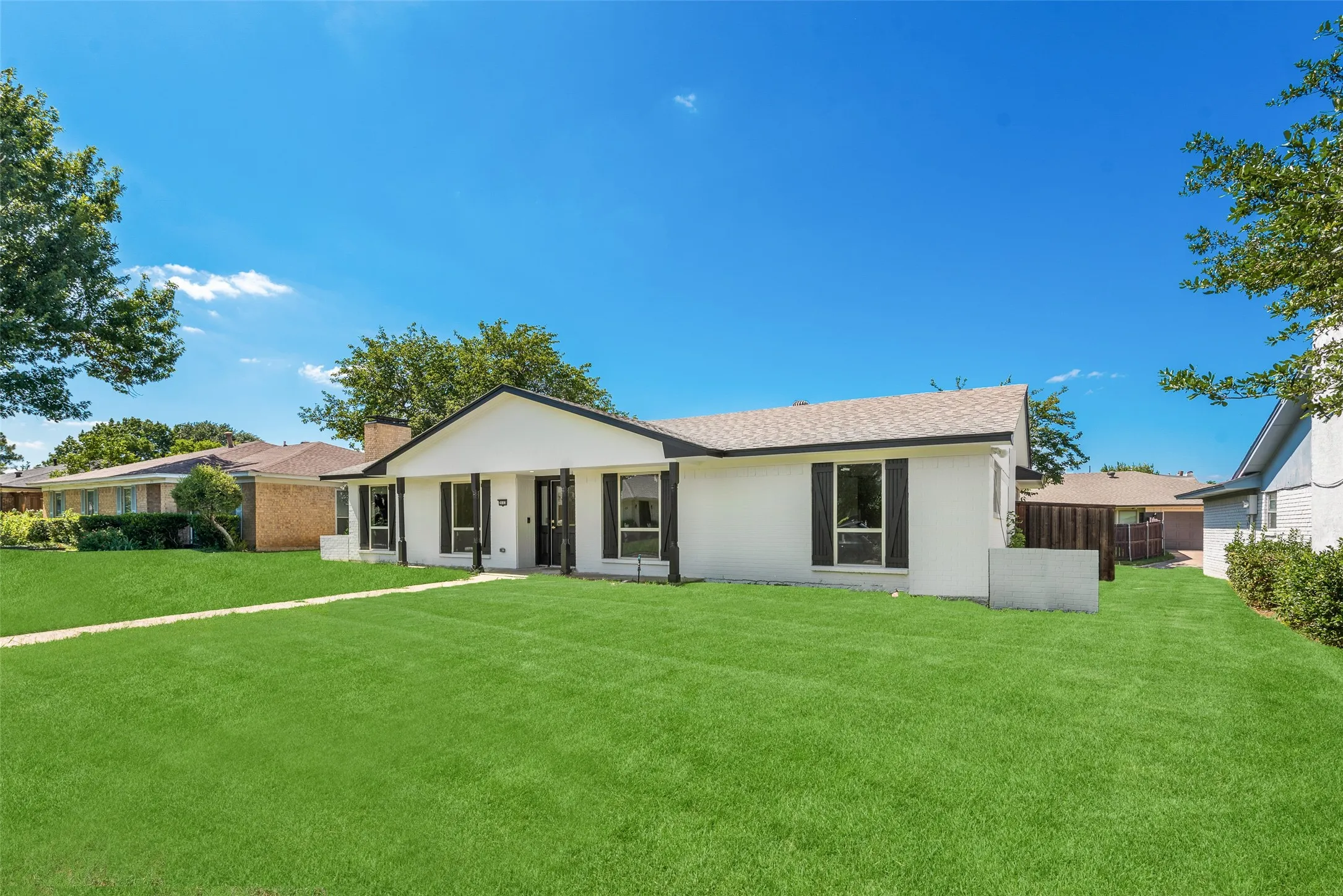 Ranch-style exterior featuring a white facade, black window shutters, a dark shingle roof, and a prominent front lawn with a concrete walkway
