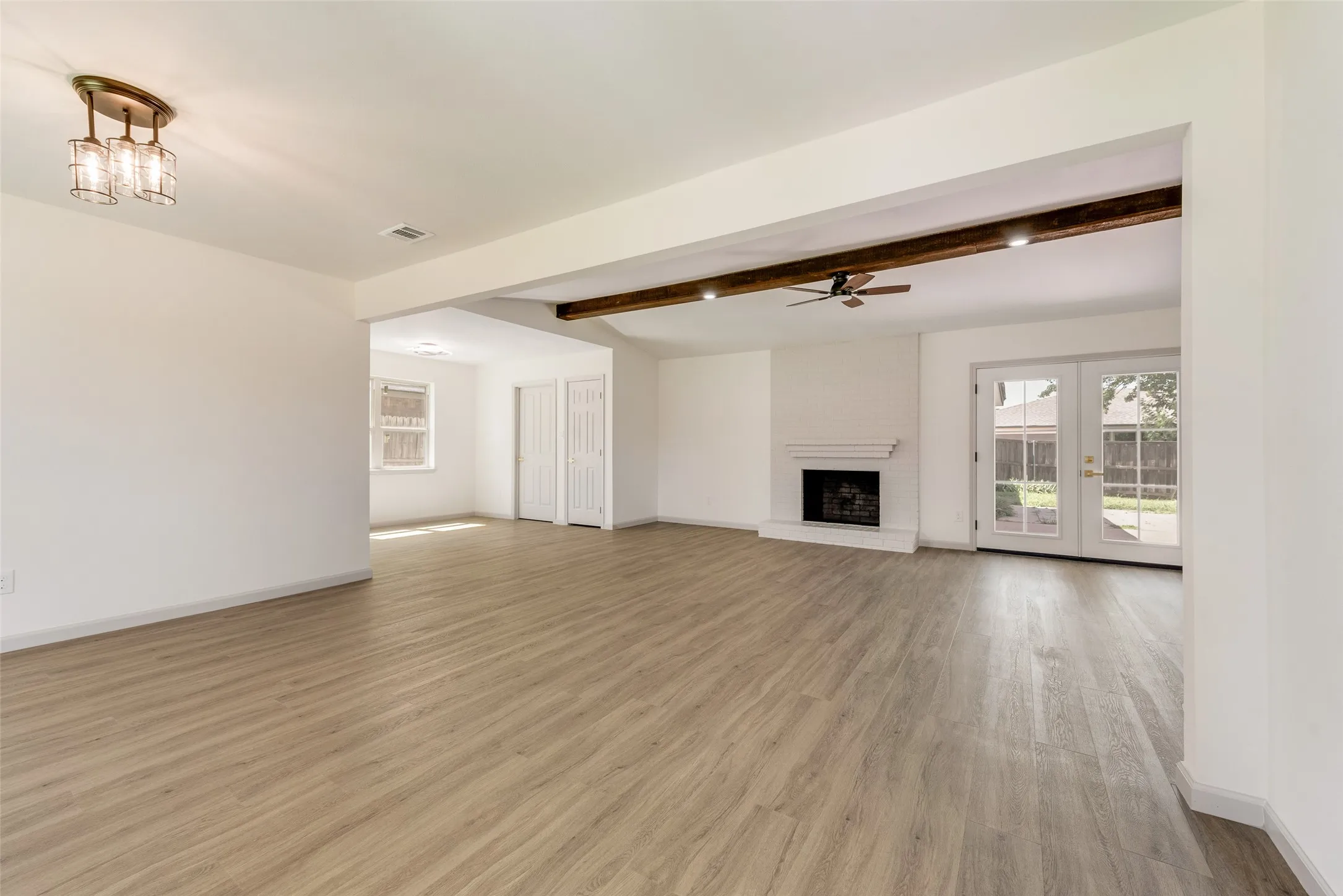 Open-concept living area featuring wood-finish flooring, a white brick fireplace with a mantel, exposed wood beams with recessed lighting, and double French doors opening to the exterior