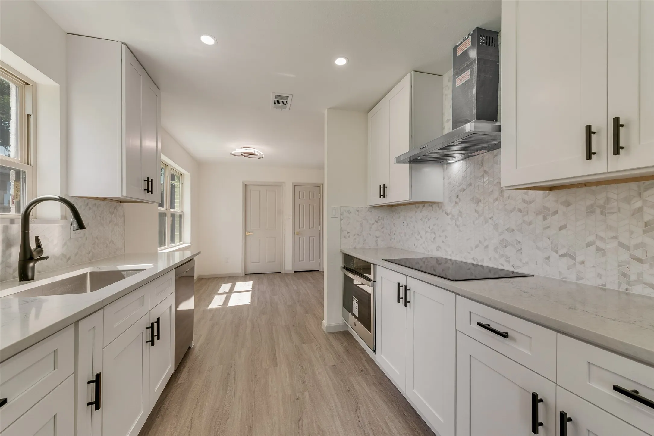 Galley kitchen featuring white shaker cabinetry, light-toned countertops, a geometric tile backsplash, stainless steel appliances, and wood-finish flooring