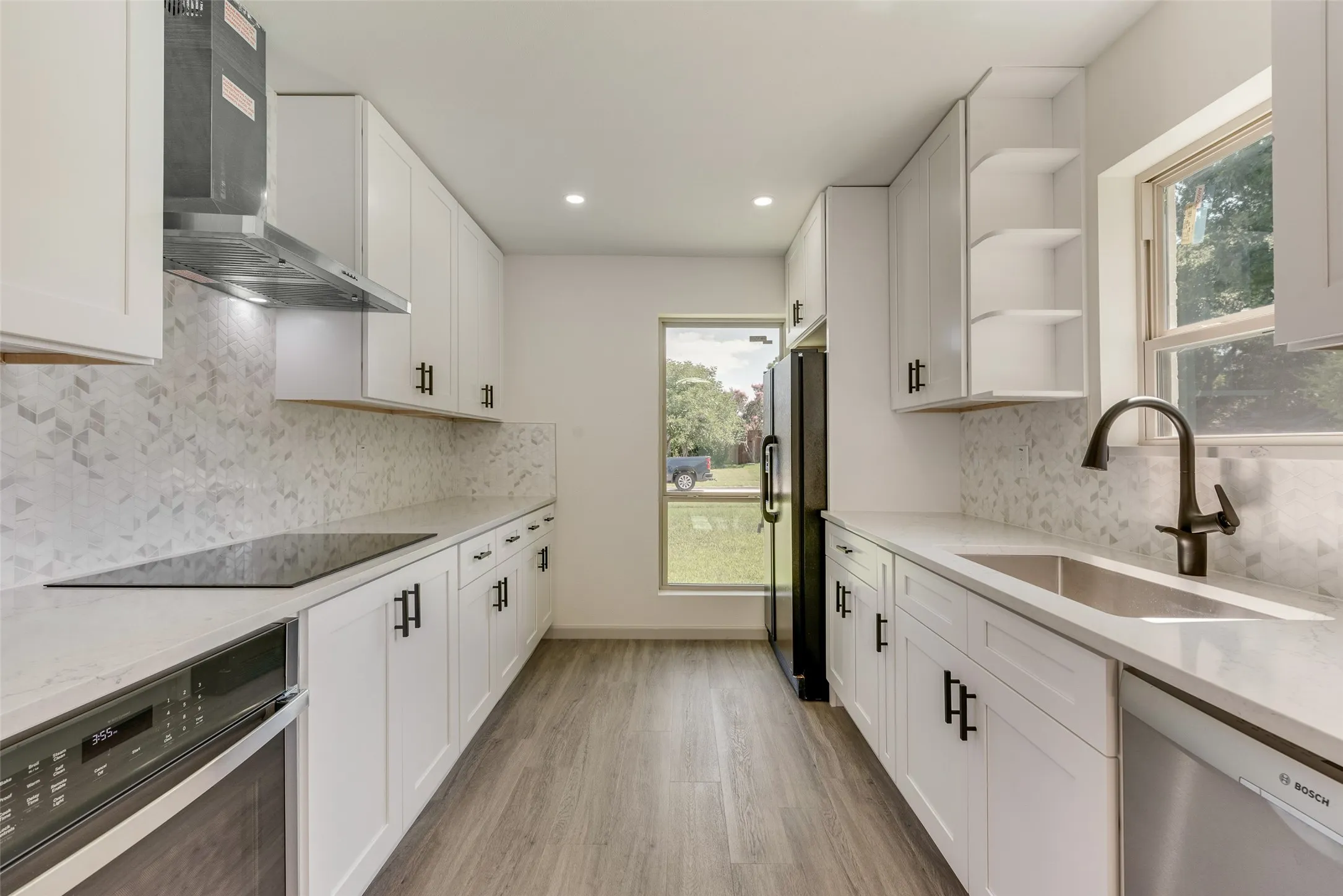 Galley kitchen featuring white cabinetry, light-toned countertops, a patterned backsplash, wood-finish flooring, and a stainless steel sink with a matte black faucet