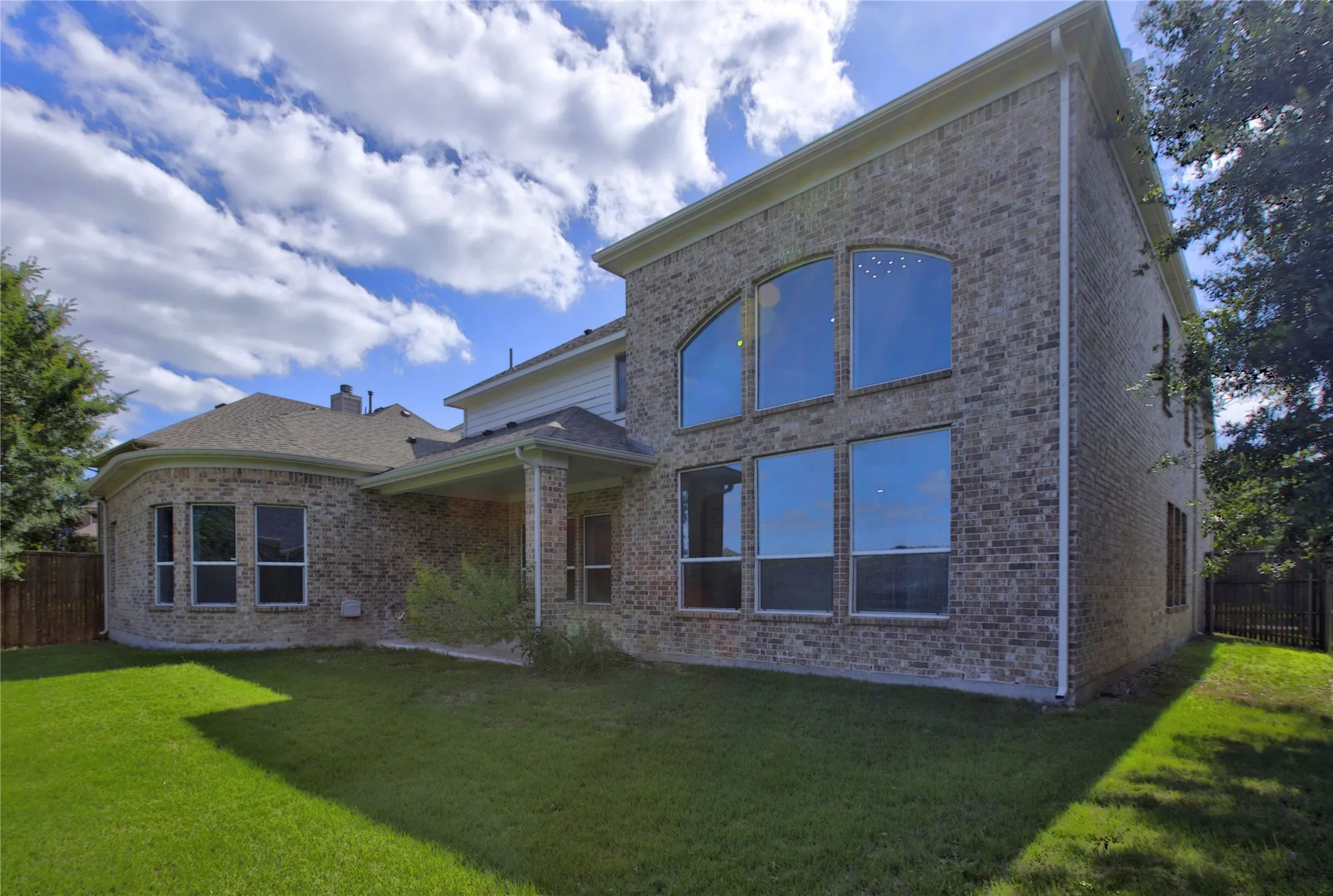 Expansive backyard featuring a manicured lawn, brick exterior, arched windows, covered patio, and privacy fencing