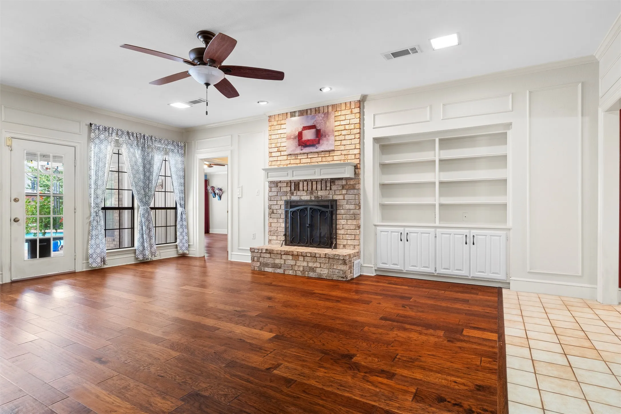 Spacious living area featuring wood-finish flooring, a brick fireplace with a mantel, built-in shelving, a ceiling fan, and French doors