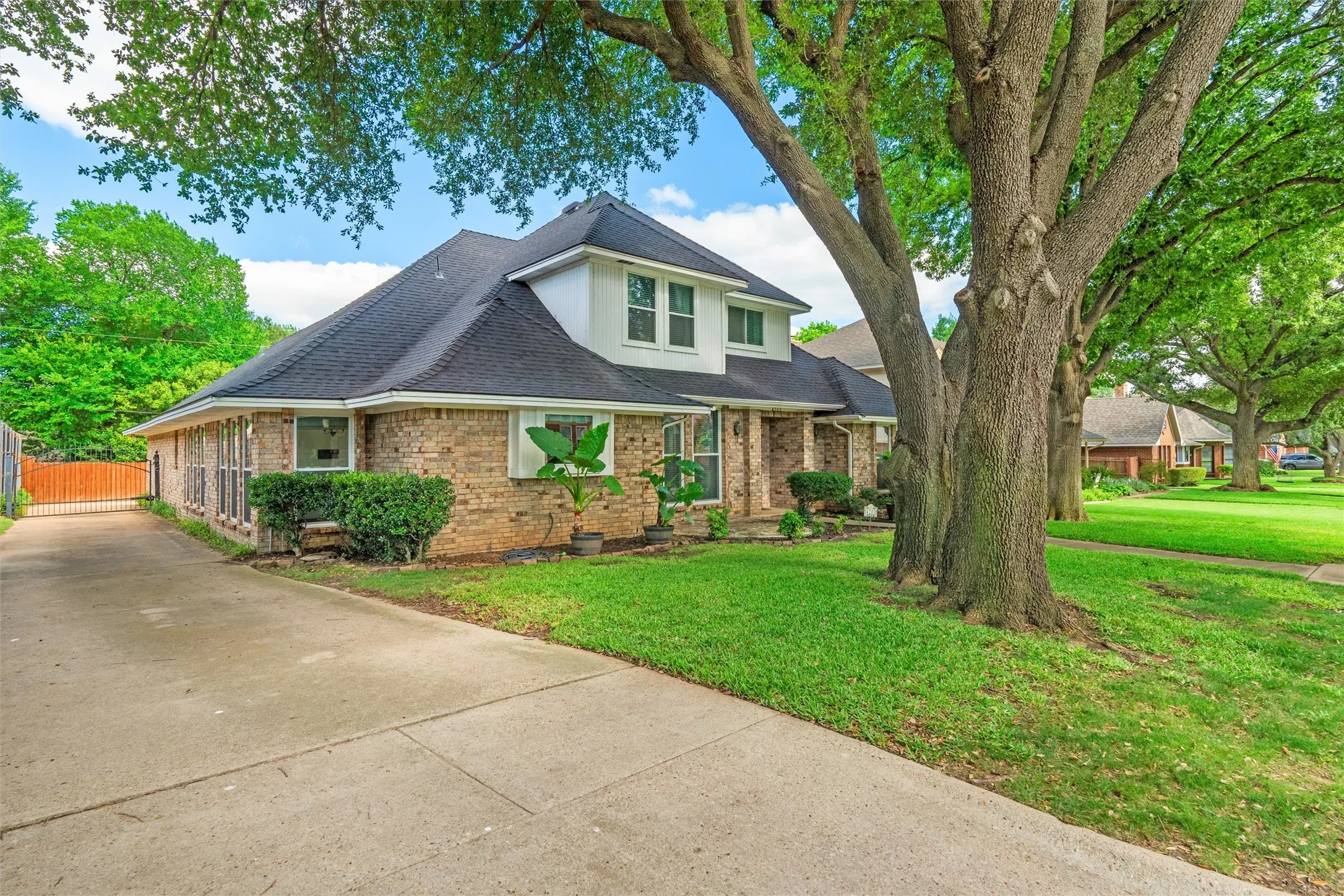 Brick residence featuring a gabled roofline, mature trees, a manicured lawn, an extended concrete driveway, and a gated side access