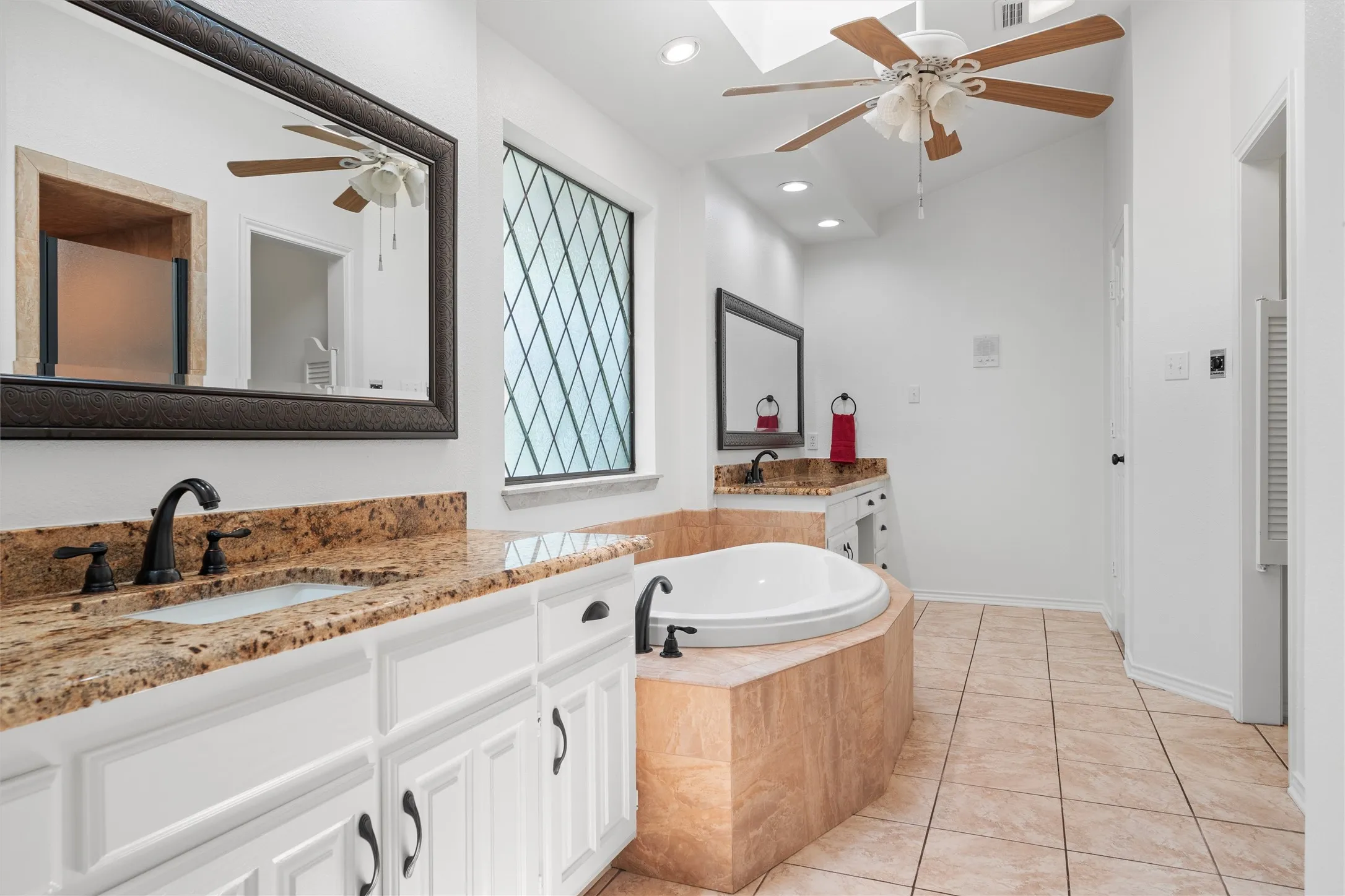 Spacious bathroom featuring a built-in soaking tub, dual vanity with granite-finish countertops, tiled flooring, and a decorative window