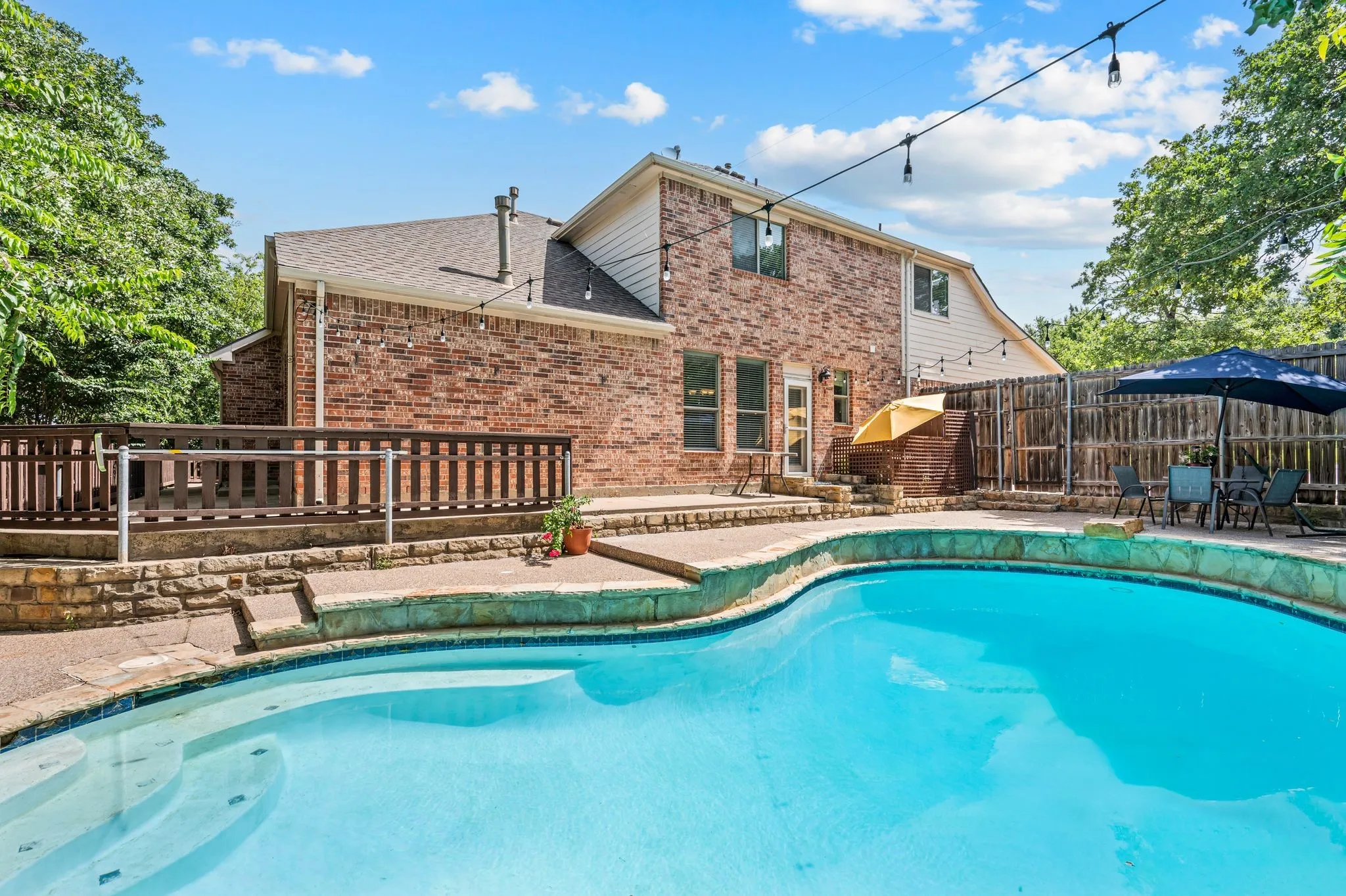 Curved inground pool with stone coping, a brick exterior home, and an elevated wood deck