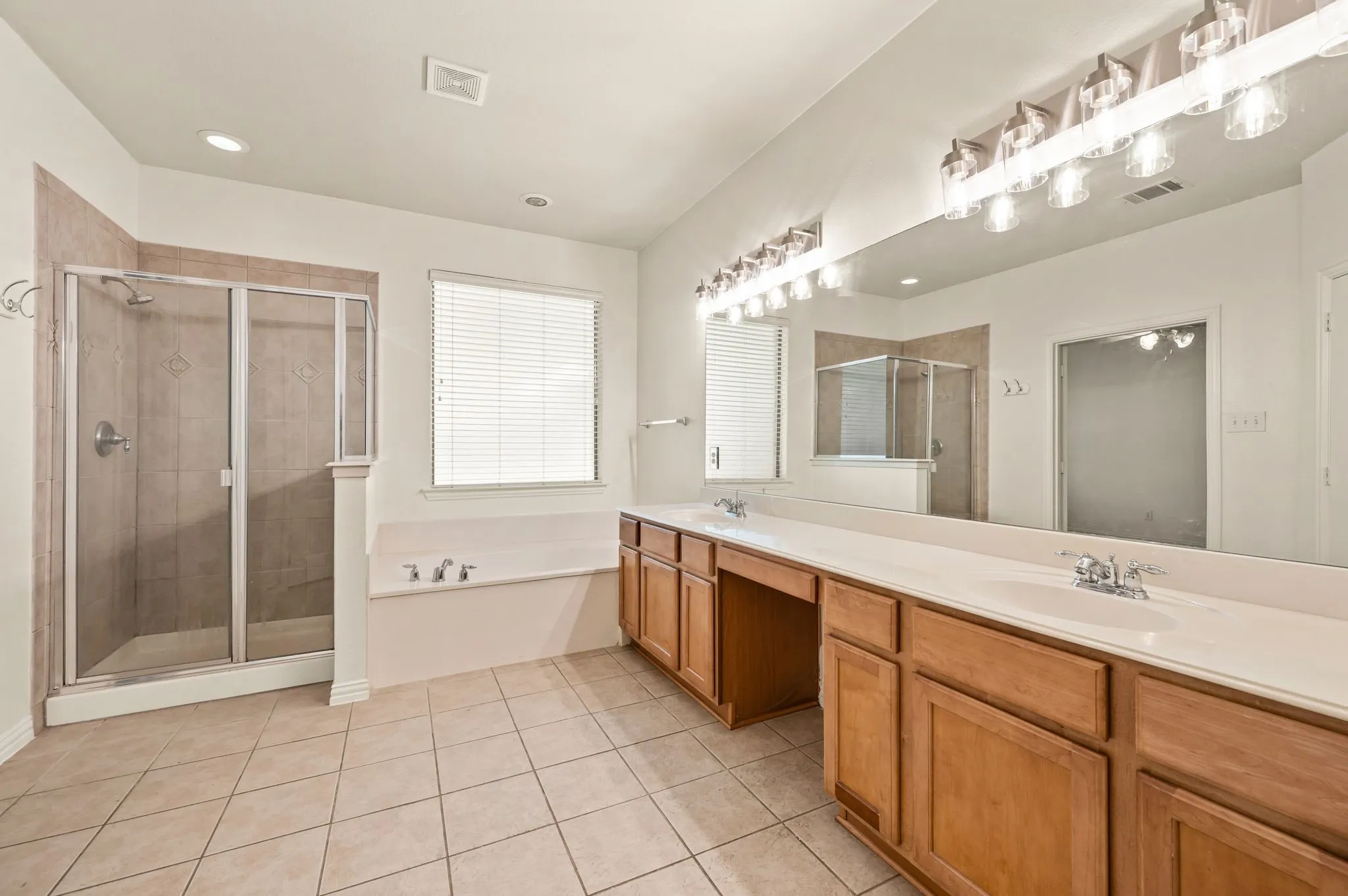 Spacious bathroom featuring twin wood-finish vanities with integrated sinks, a large vanity mirror with overhead lighting, and a glass-enclosed shower with tile surround