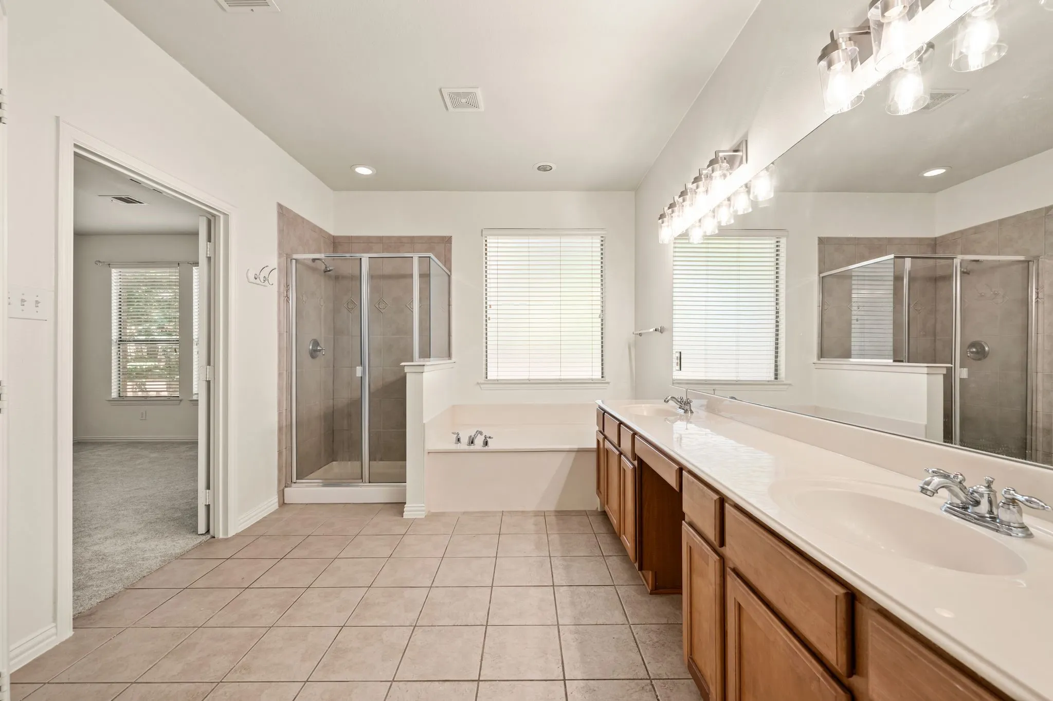 Spacious bathroom featuring dual vanities with integrated sinks, a wide mirror with overhead lighting, a glass-enclosed shower, a built-in bathtub, and tile flooring