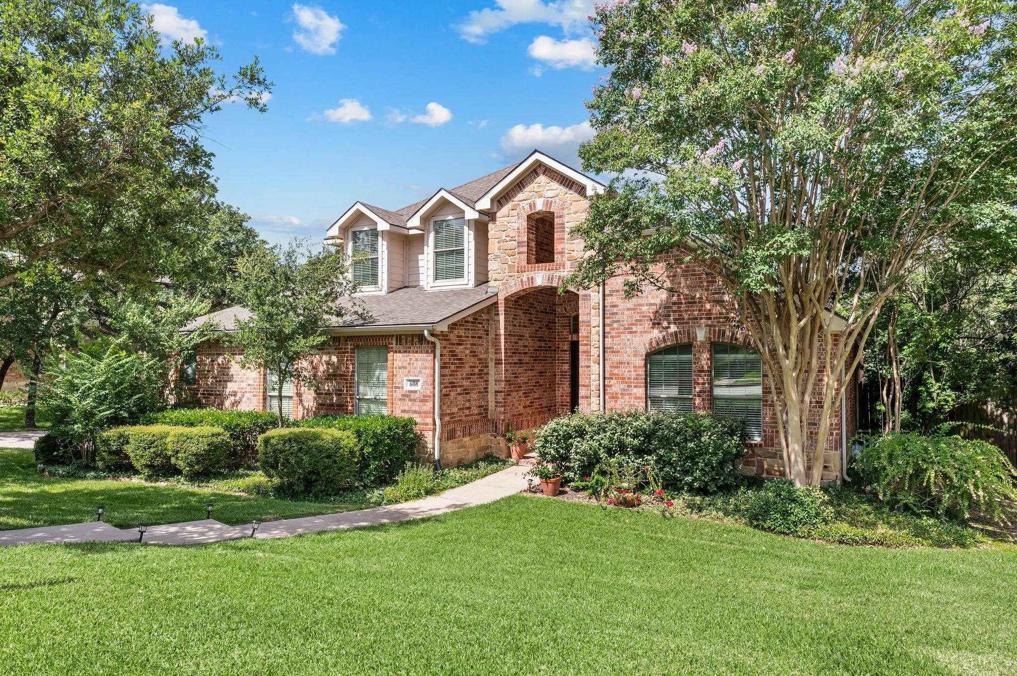 Traditional brick facade residence with a prominent arched entry, multiple dormer windows, a concrete walkway, established landscaping, and a manicured lawn