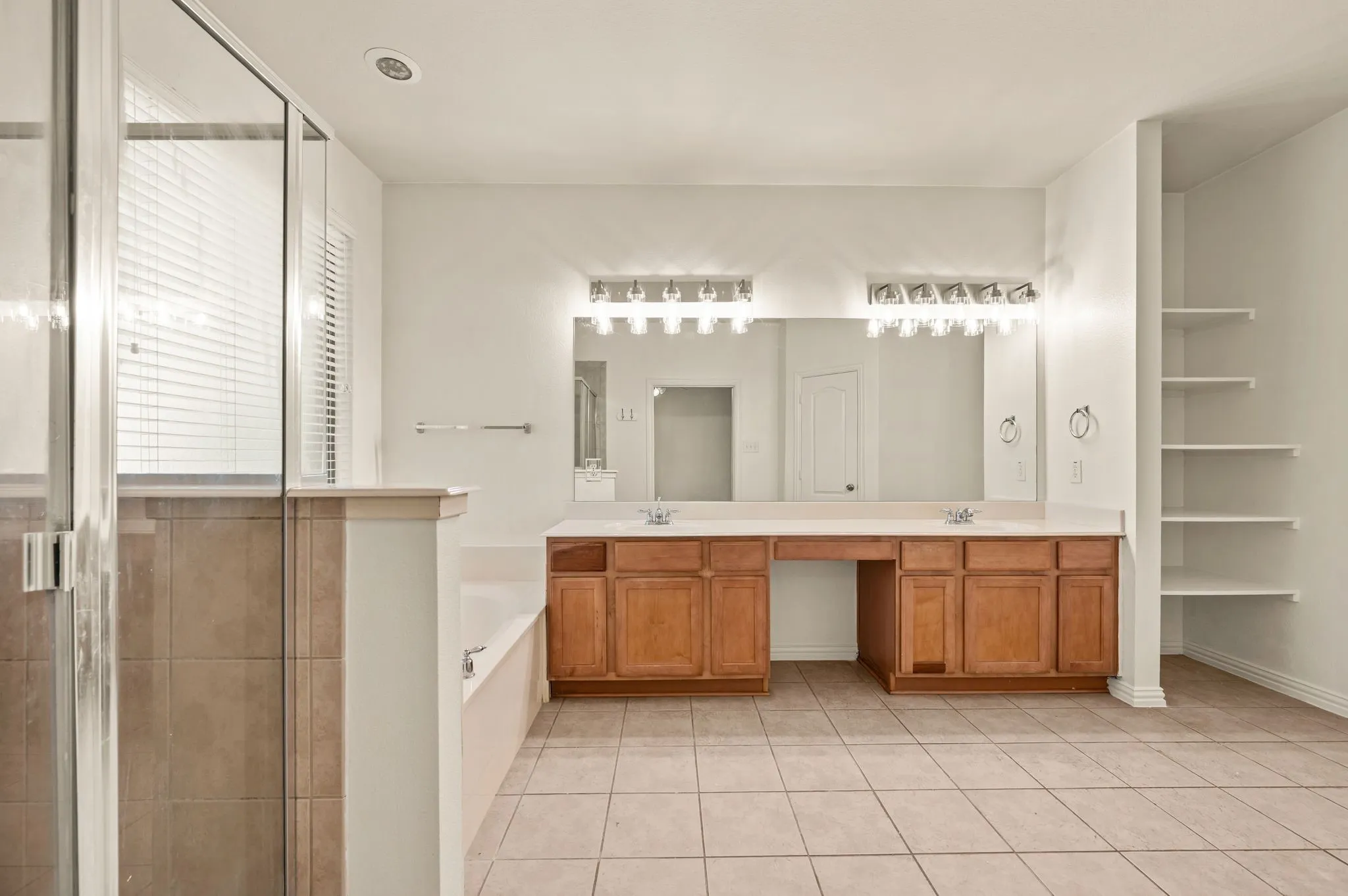 Spacious bathroom featuring a dual vanity with wood cabinetry, a large soaking tub, a separate glass-enclosed shower with tile surround, and built-in shelving
