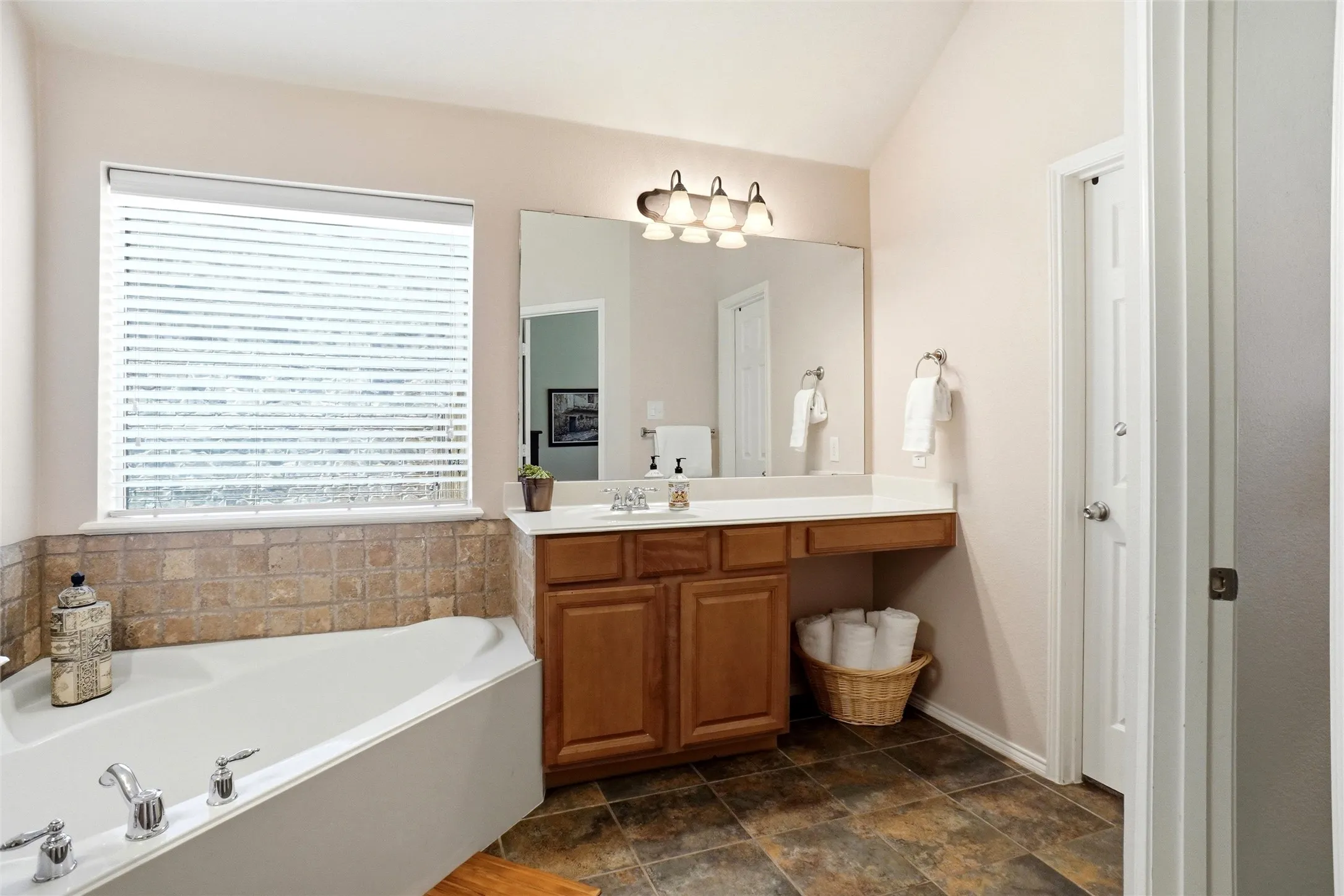 Bathroom featuring a corner soaking tub with tile surround, a single vanity with wood cabinetry, a large mirror, and a window with blinds