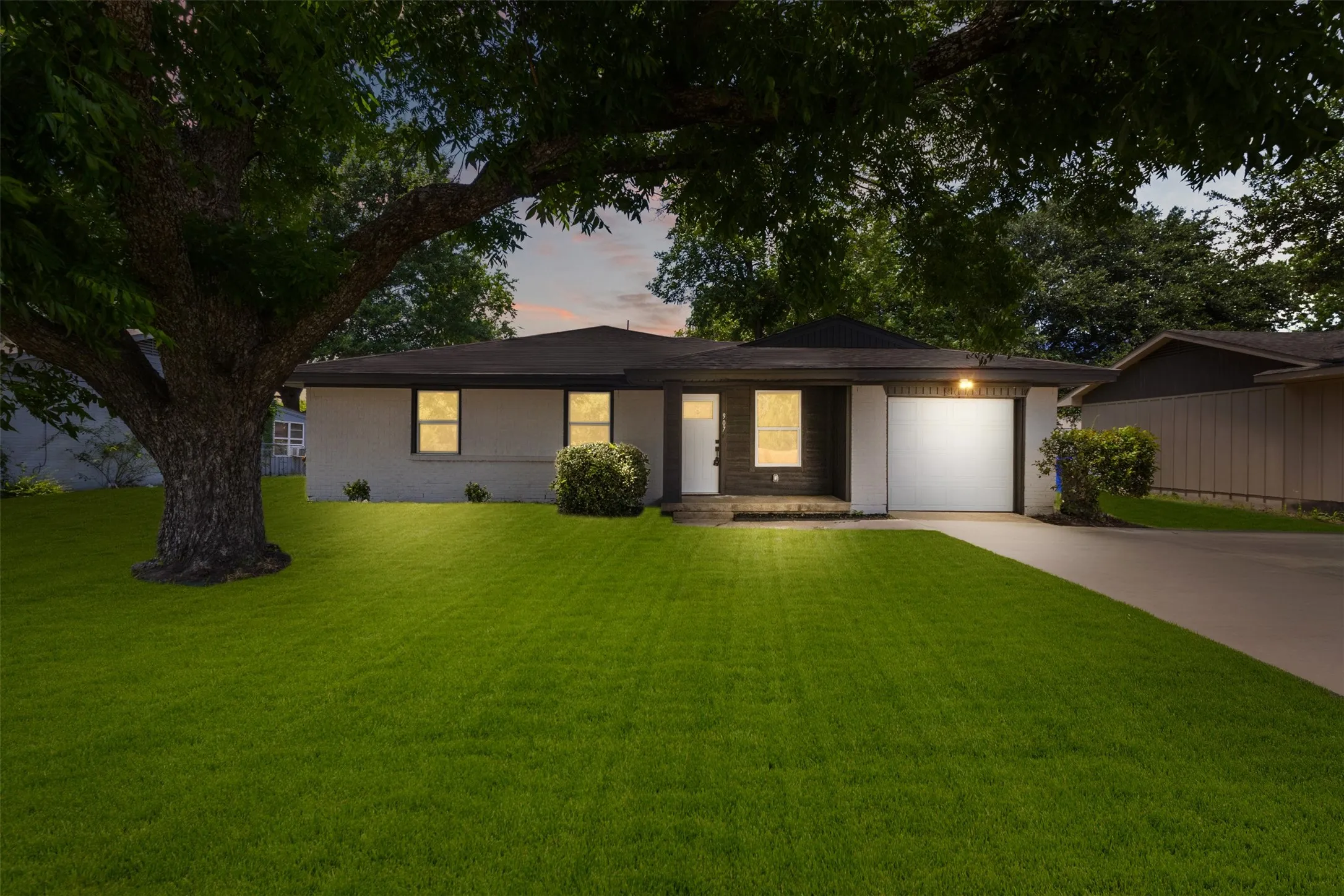 Single-story residence featuring a white brick exterior, a dark shingle roof, and a prominent mature tree providing ample shade