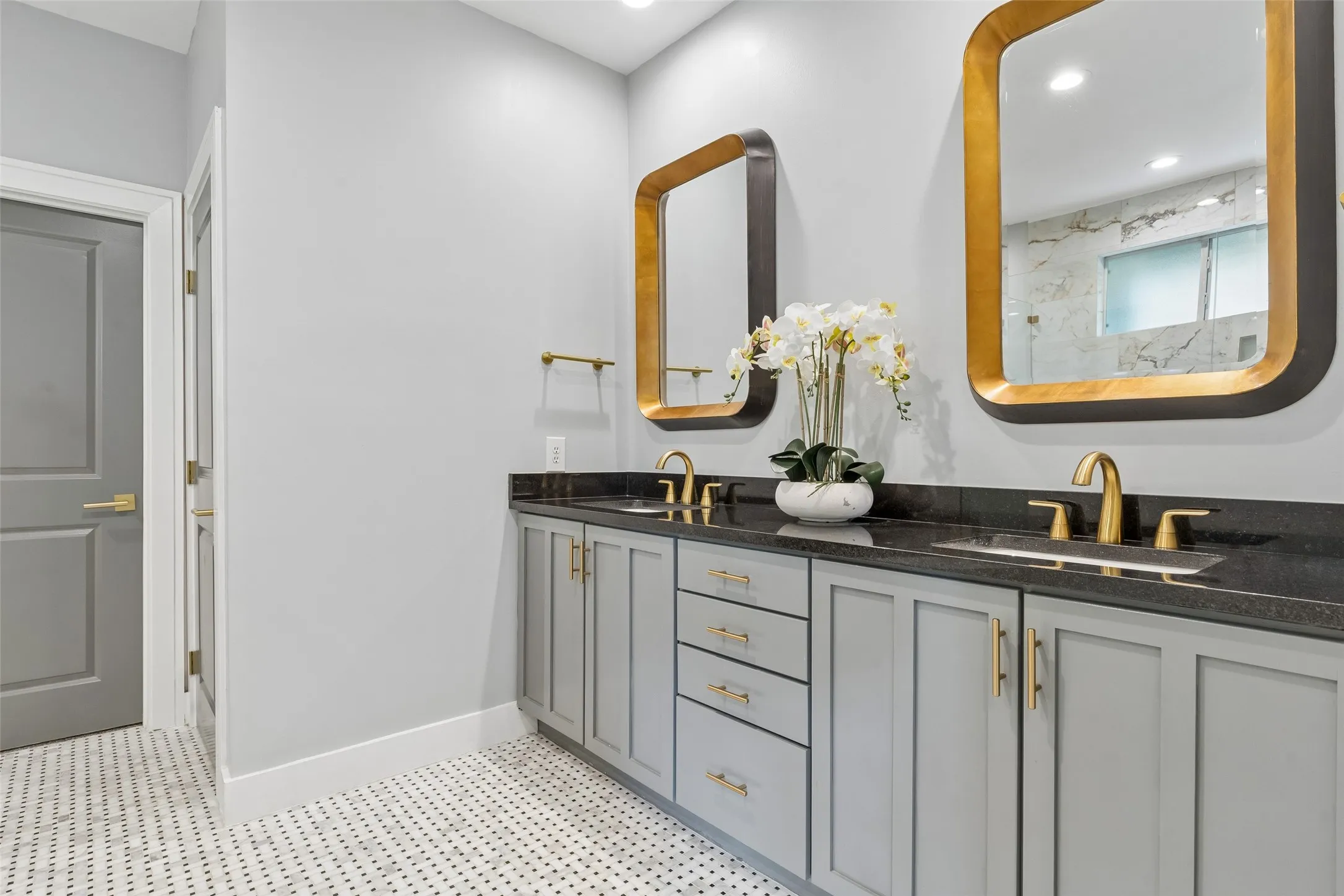 Dual vanity featuring gold-toned fixtures, black countertops, and light gray cabinetry with gold hardware