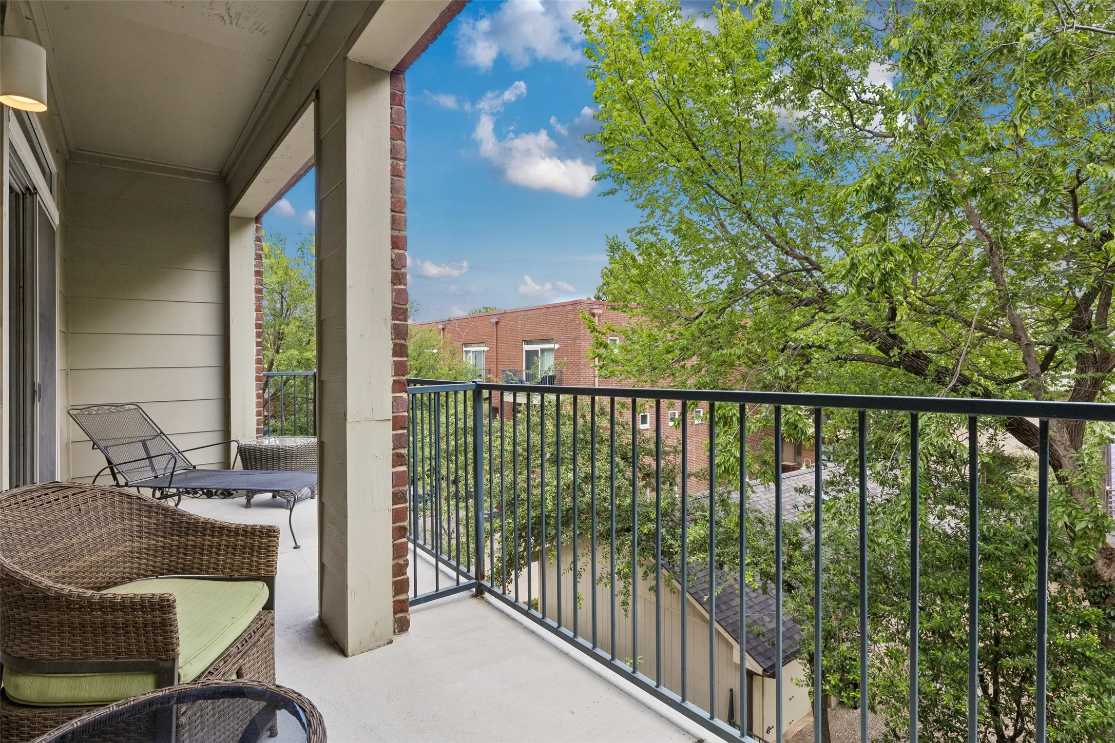 Spacious private balcony featuring a concrete floor, brick and siding exterior, black metal railing, and an overhead light fixture