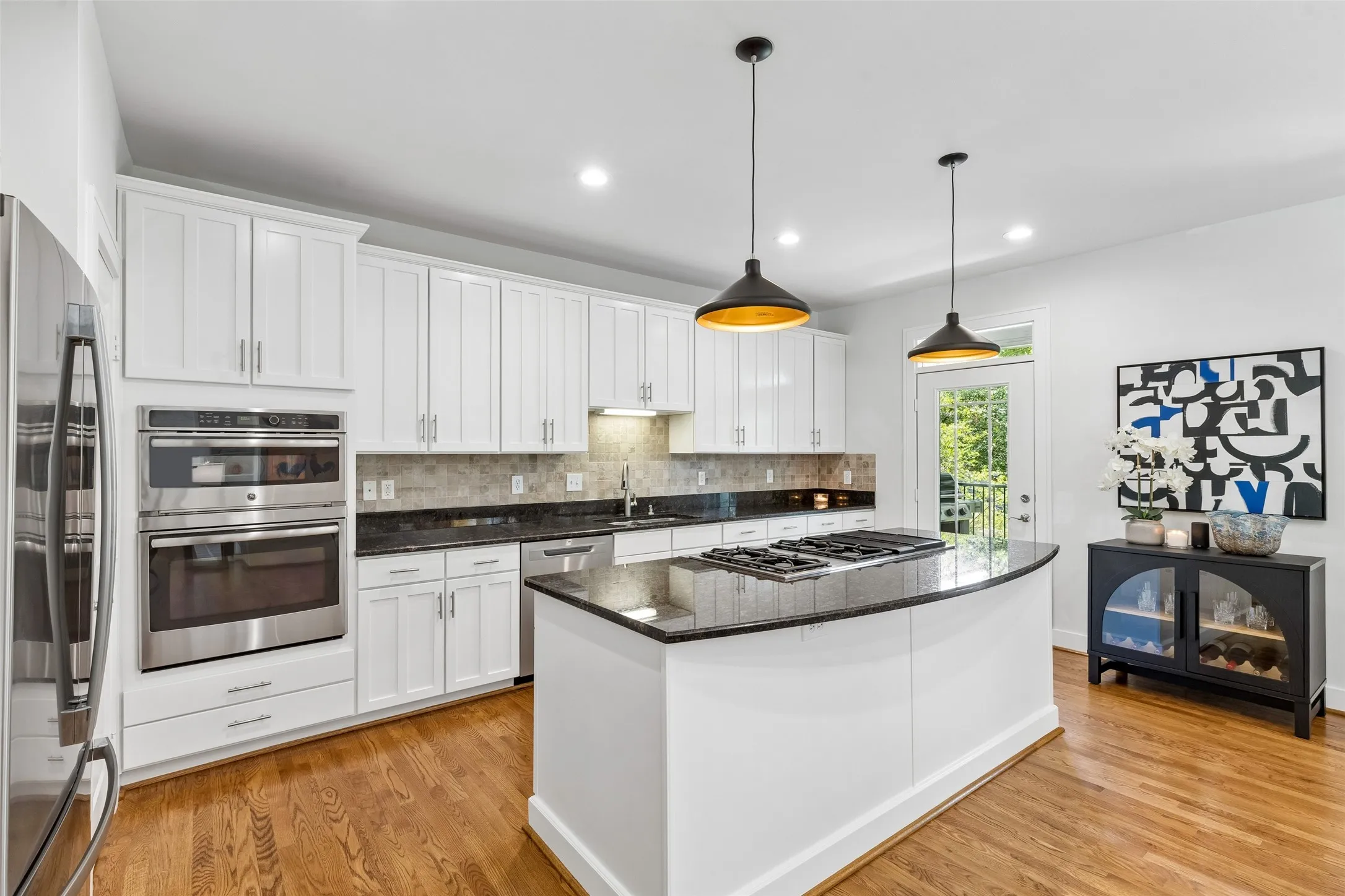 Gourmet kitchen featuring a curved island with integrated cooktop, white shaker cabinetry, stainless steel appliances, dark stone countertops, and wood-finish flooring