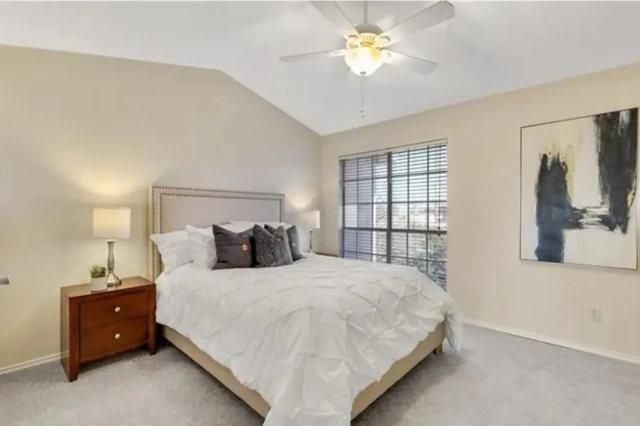 Vaulted ceiling bedroom featuring light beige walls, a neutral carpet, a large window with horizontal blinds, and a ceiling fan with integrated lighting